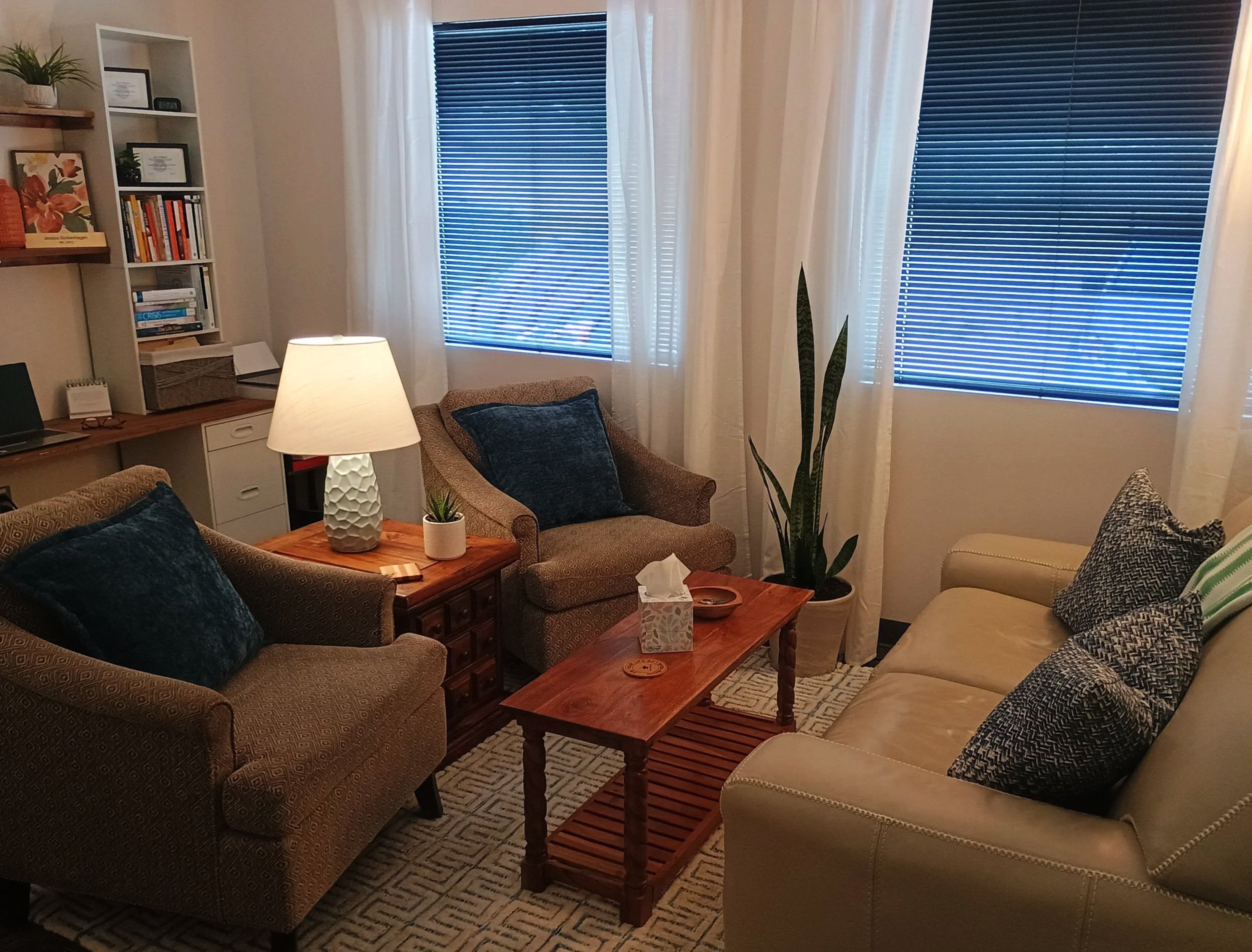 A peaceful therapy room with two large windows, two cozy armchairs, a tan leather loveseat, blue throw pillows, a wooden coffee table, a floor plant, a lamp, and a bookshelf.
