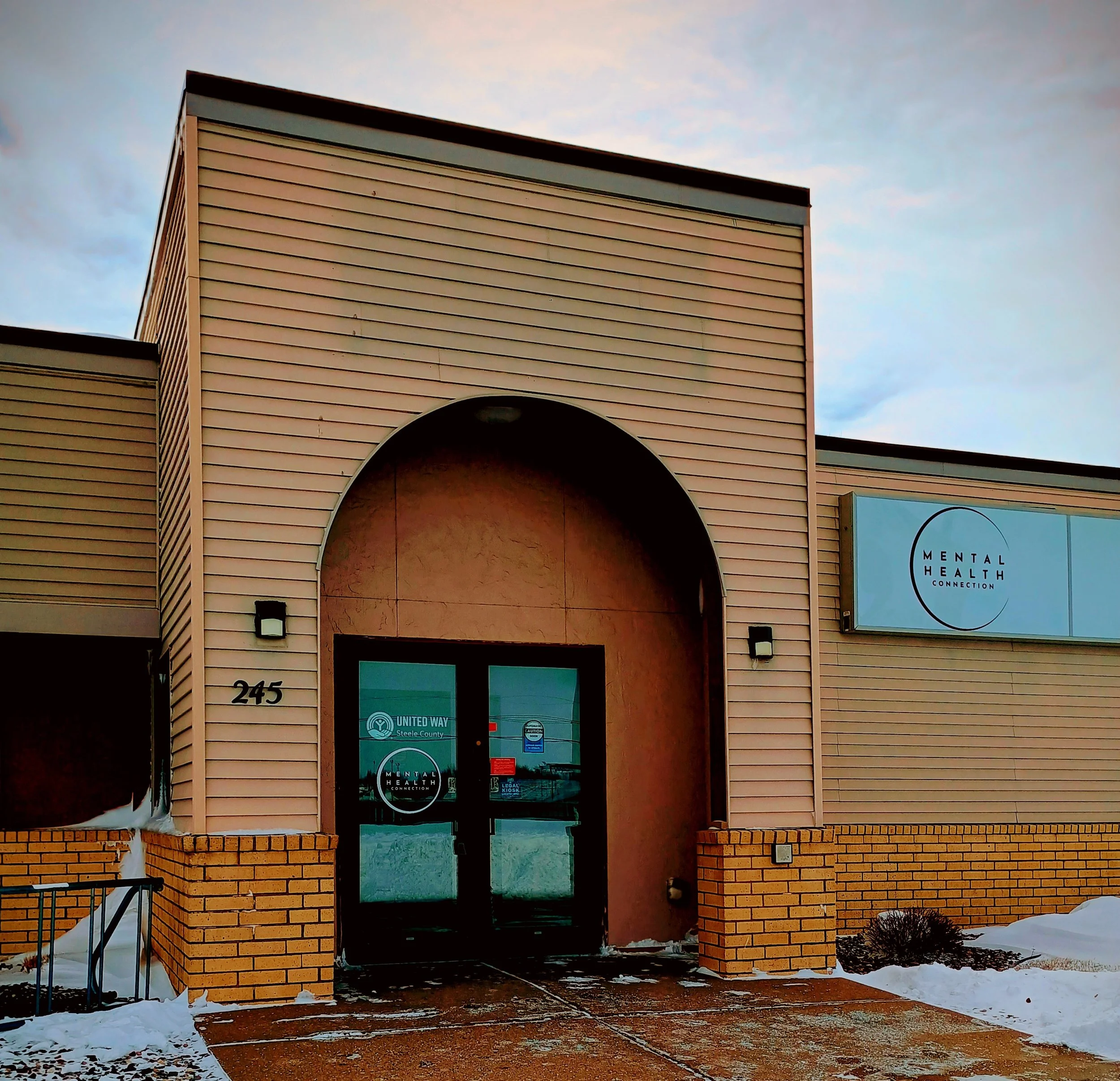 Entrance of a mental health clinic with glass doors, beige siding, brick accents, and a sign reading 'Mental Health Connection' outside.