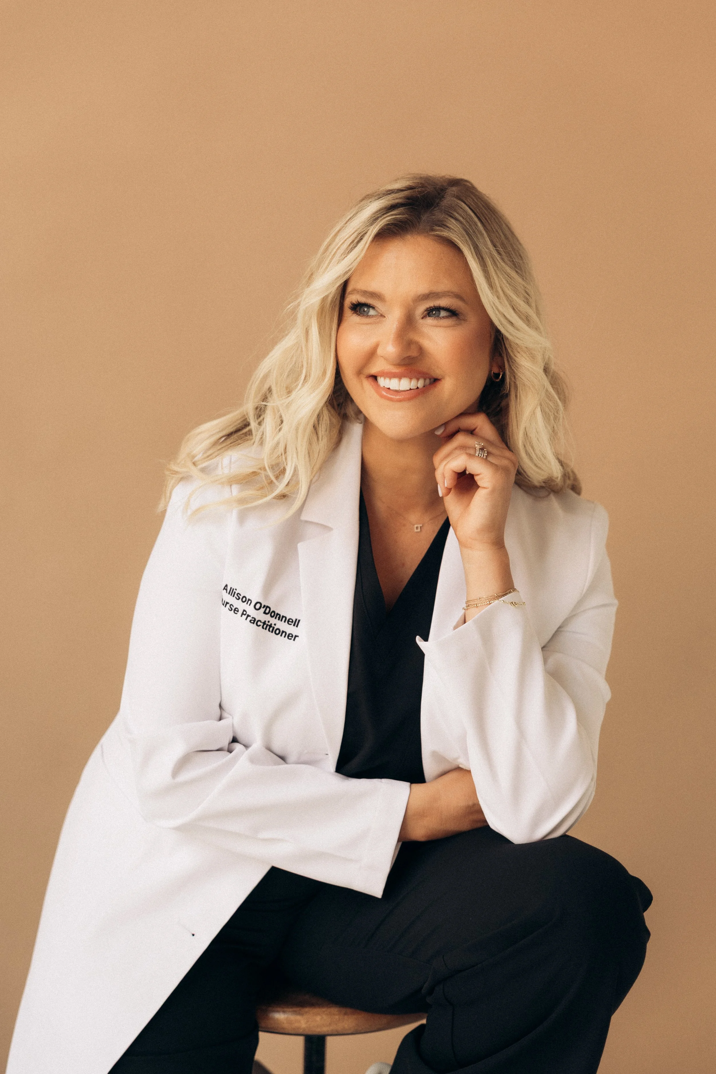 A smiling woman with blonde hair wearing a white coat with her name and title, sitting on a stool against a beige background.