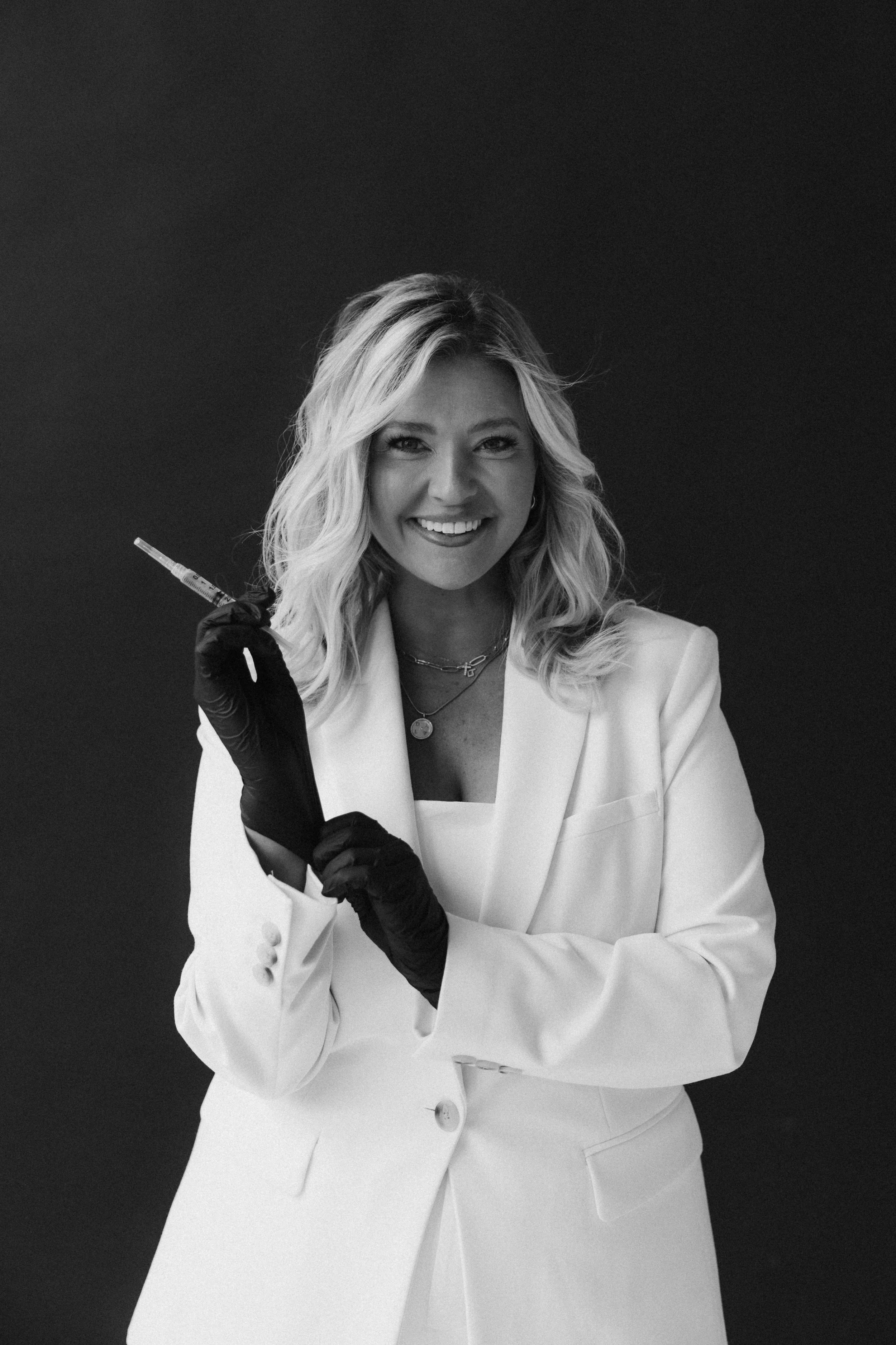 A woman with blonde hair smiling, dressed in a white blazer, holding a syringe, wearing black gloves, against a dark background.