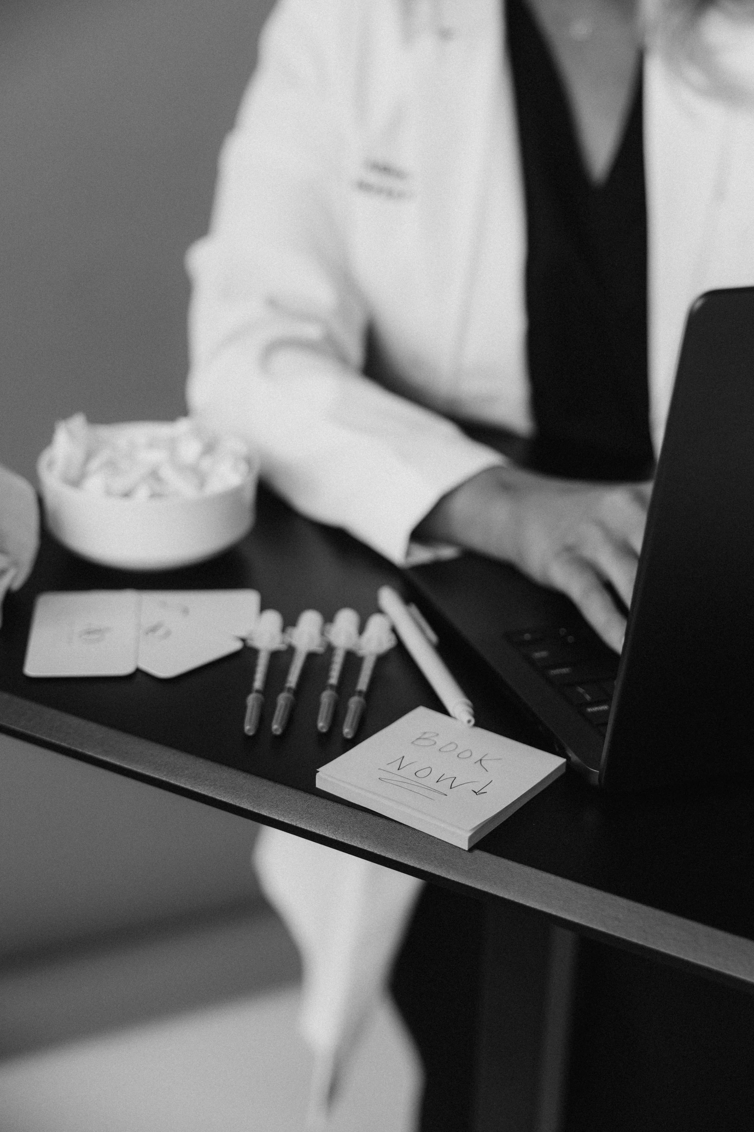 Medical professional wearing a white coat working on a laptop at a desk with a notepad, darts, and a bowl of crumpled paper.