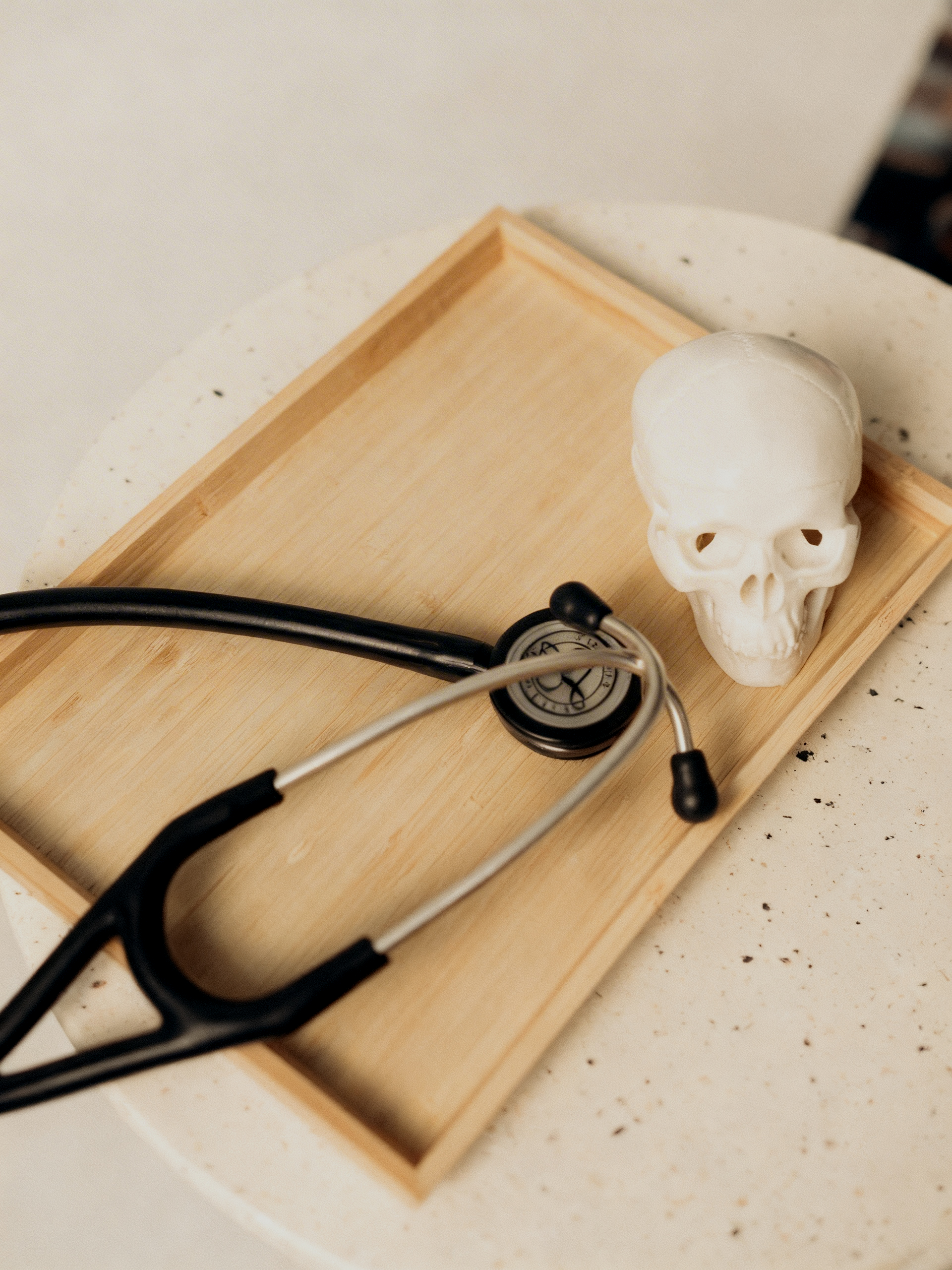 A small wooden tray on a white speckled table holds a white skull, a stethoscope with black and silver parts, and a small round clock.
