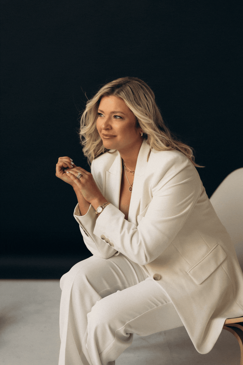 A woman wearing a white suit sitting on a chair against a dark background, with blonde hair, smiling gently and looking to her left.
