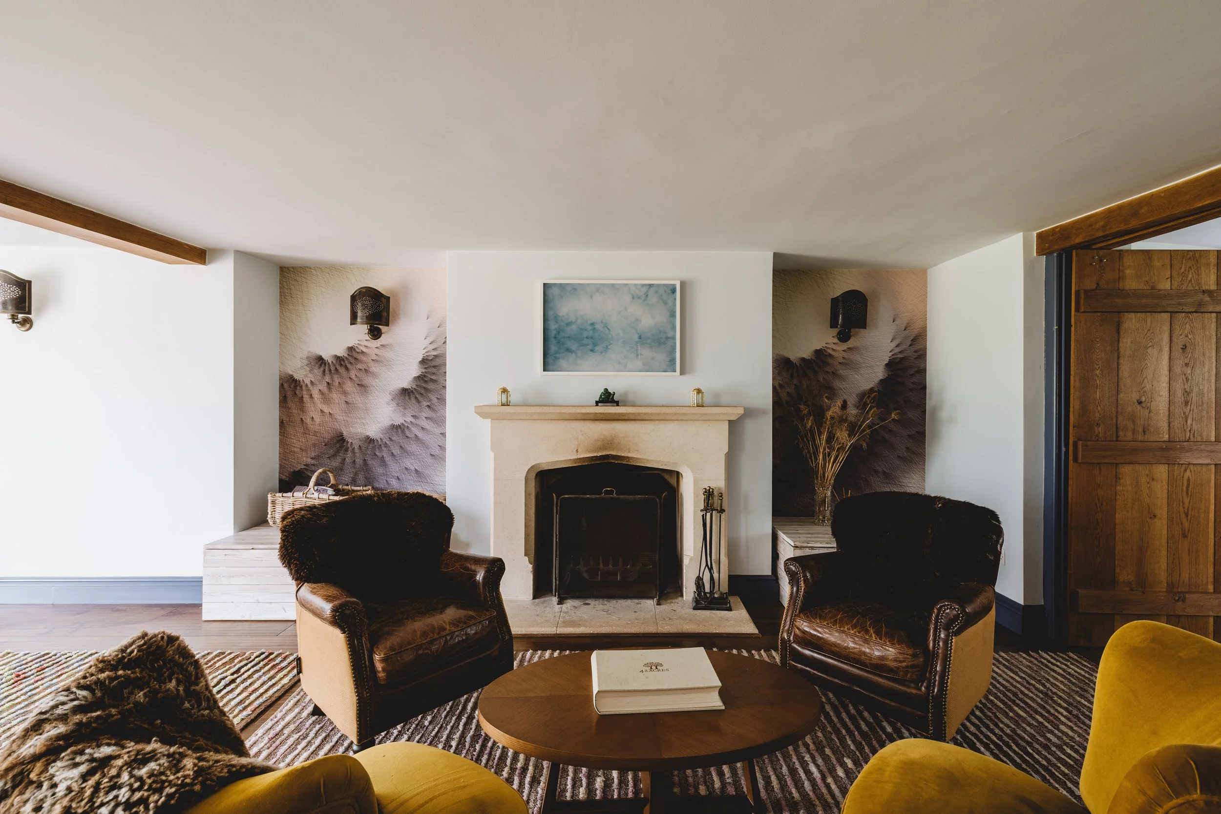 Cozy living room with a fireplace, two brown leather armchairs, a wooden coffee table with a book, and yellow upholstered furniture. Decor includes wall art, dried plants, and wall sconces.