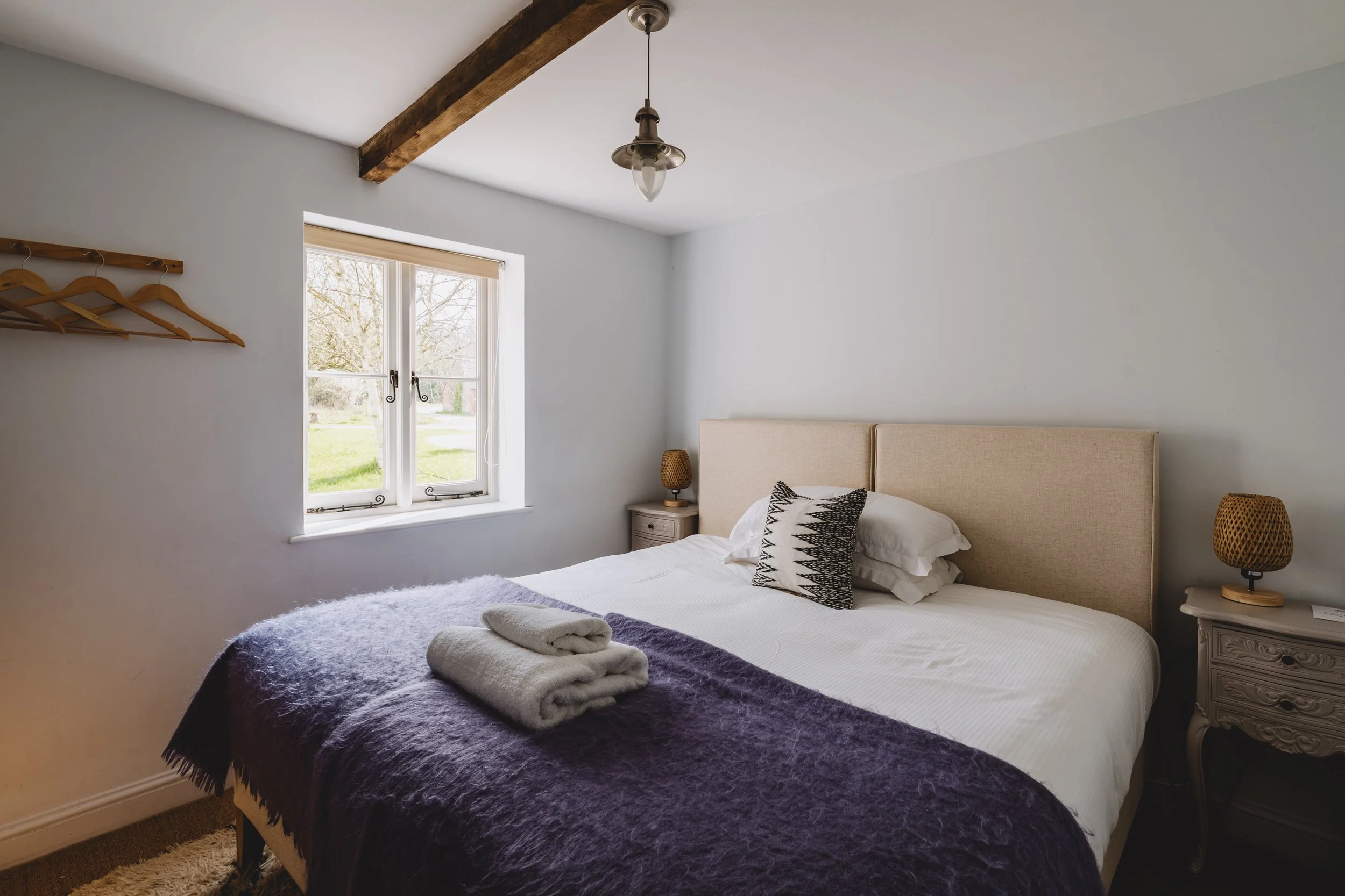 A bedroom with a large bed covered in white sheets, with folded towels placed on a purple blanket at the foot of the bed. The room has a window with a view of a grassy yard and trees, two matching nightstands with woven lamps, and a wooden beam across the ceiling.