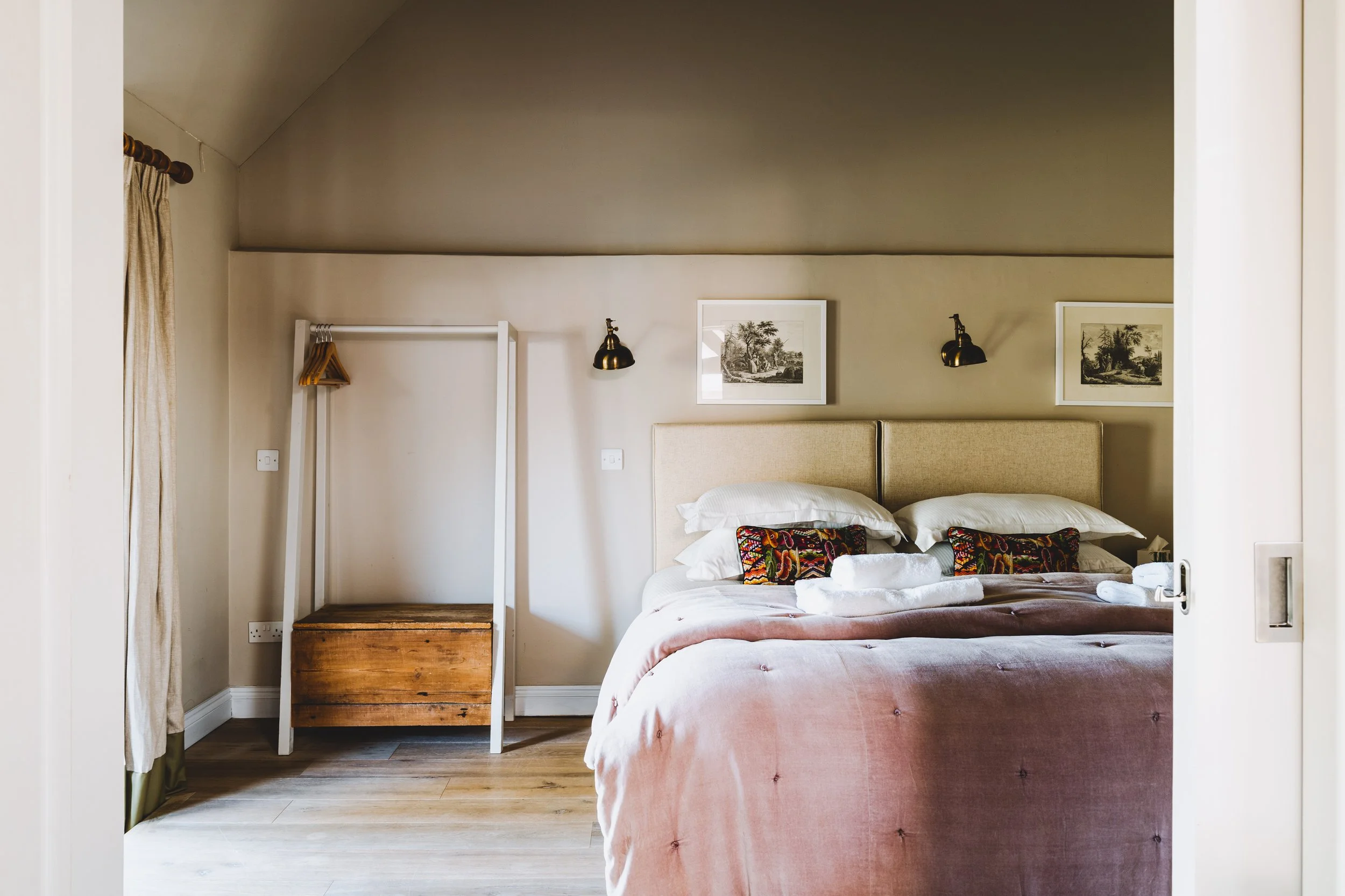 A neatly made bedroom with a large bed, beige headboard, and colorful pillows, flanked by two framed pictures on the wall, and a small wooden clothing rack on the left side of the room.