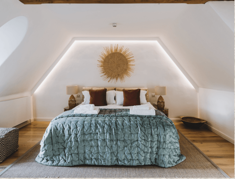 A cozy bedroom with a large bed, teal quilt, white and brown pillows, two bedside lamps, a sunburst wall decor above the bed, and wooden flooring.