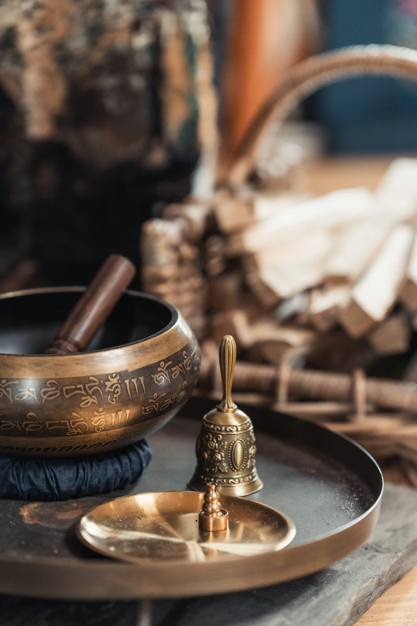 A Tibetan singing bowl with a mallet resting inside, placed on a round black cushion, on a round metal tray, accompanied by a small brass bell and a brass incense holder, with neatly stacked firewood in the background.