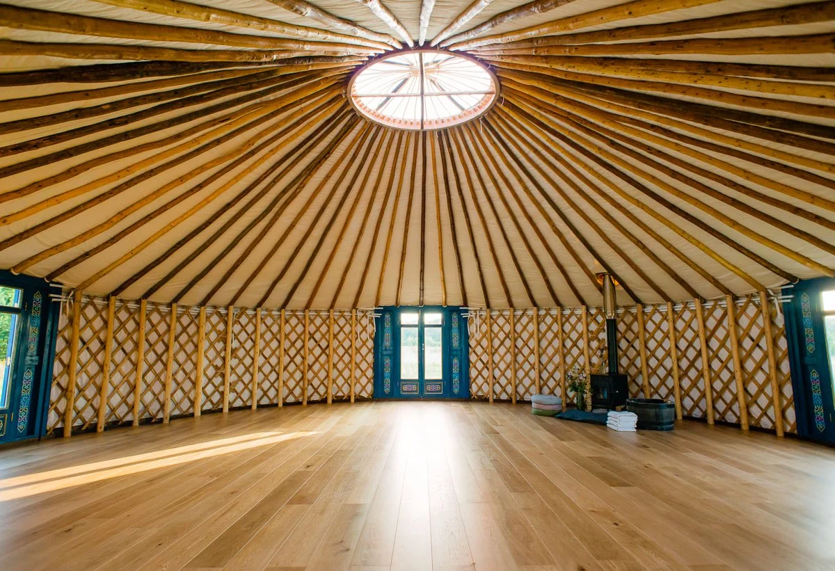 Interior of a yurt with wooden lattice walls, a circular skylight at the center of the ceiling, and a wood-burning stove in the corner