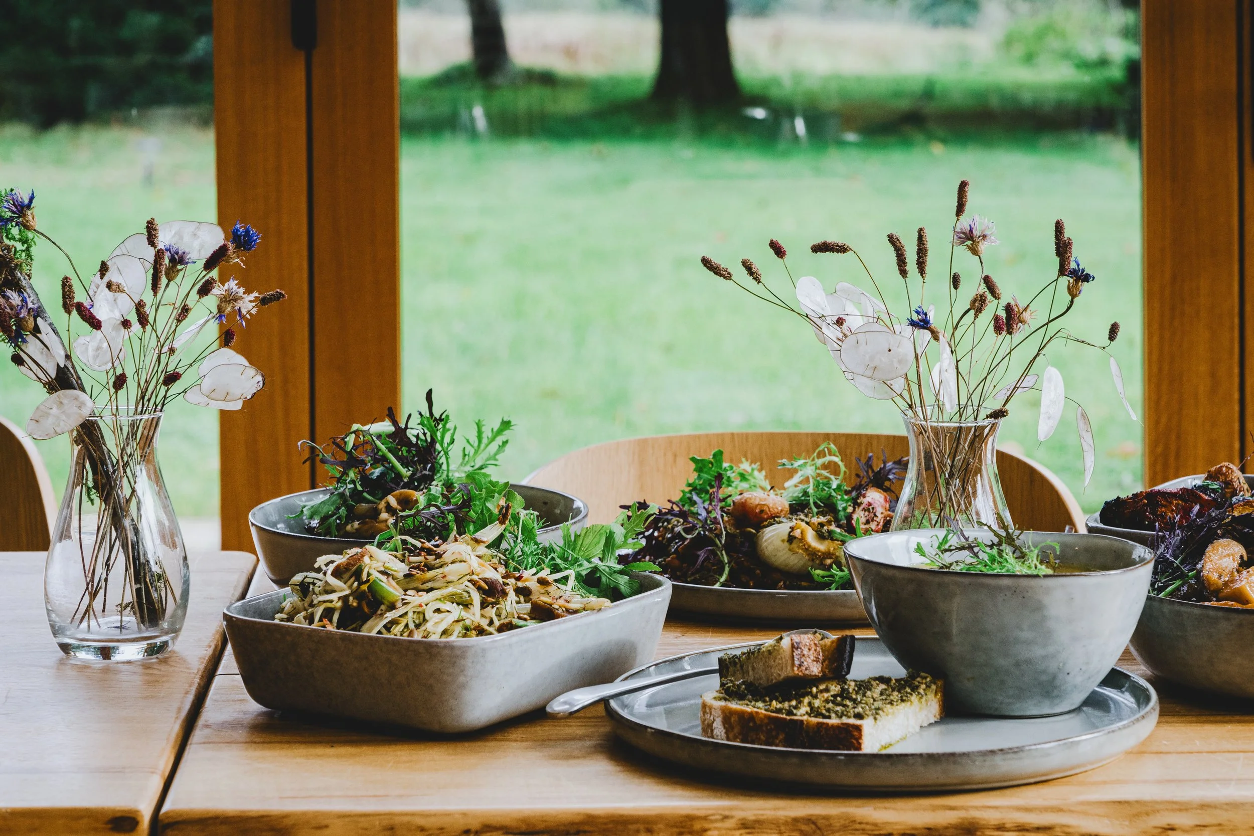 Table set with salads, bread, and flowers in vases near a window with green outdoor scenery.