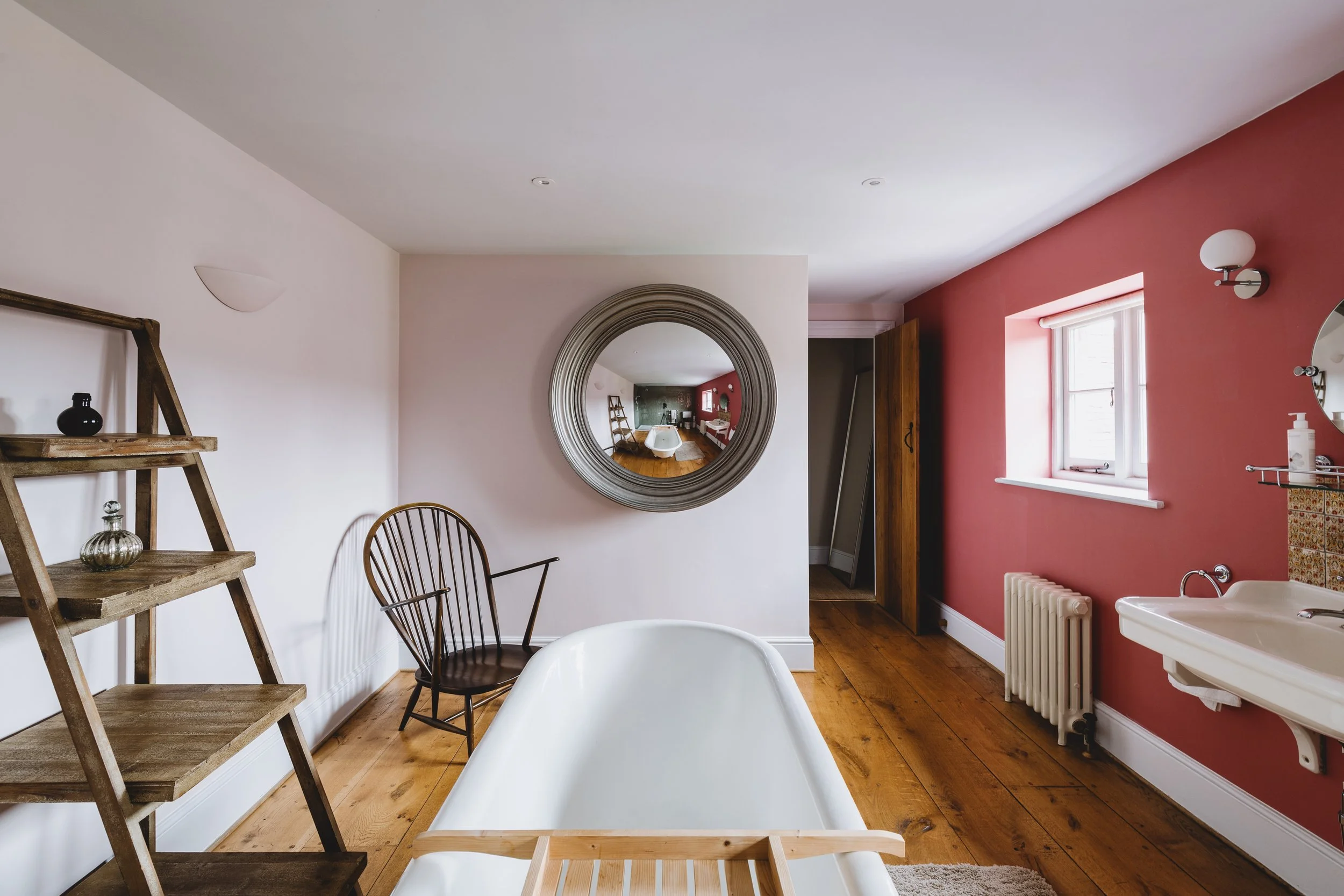 A bathroom with a white bathtub at the center, a wooden chair to the left, a wooden ladder shelf with small decorative items, a round mirror on the wall, a window with a pink wall, a radiator below the window, and a sink on the right side, with vintage-style lighting fixtures.