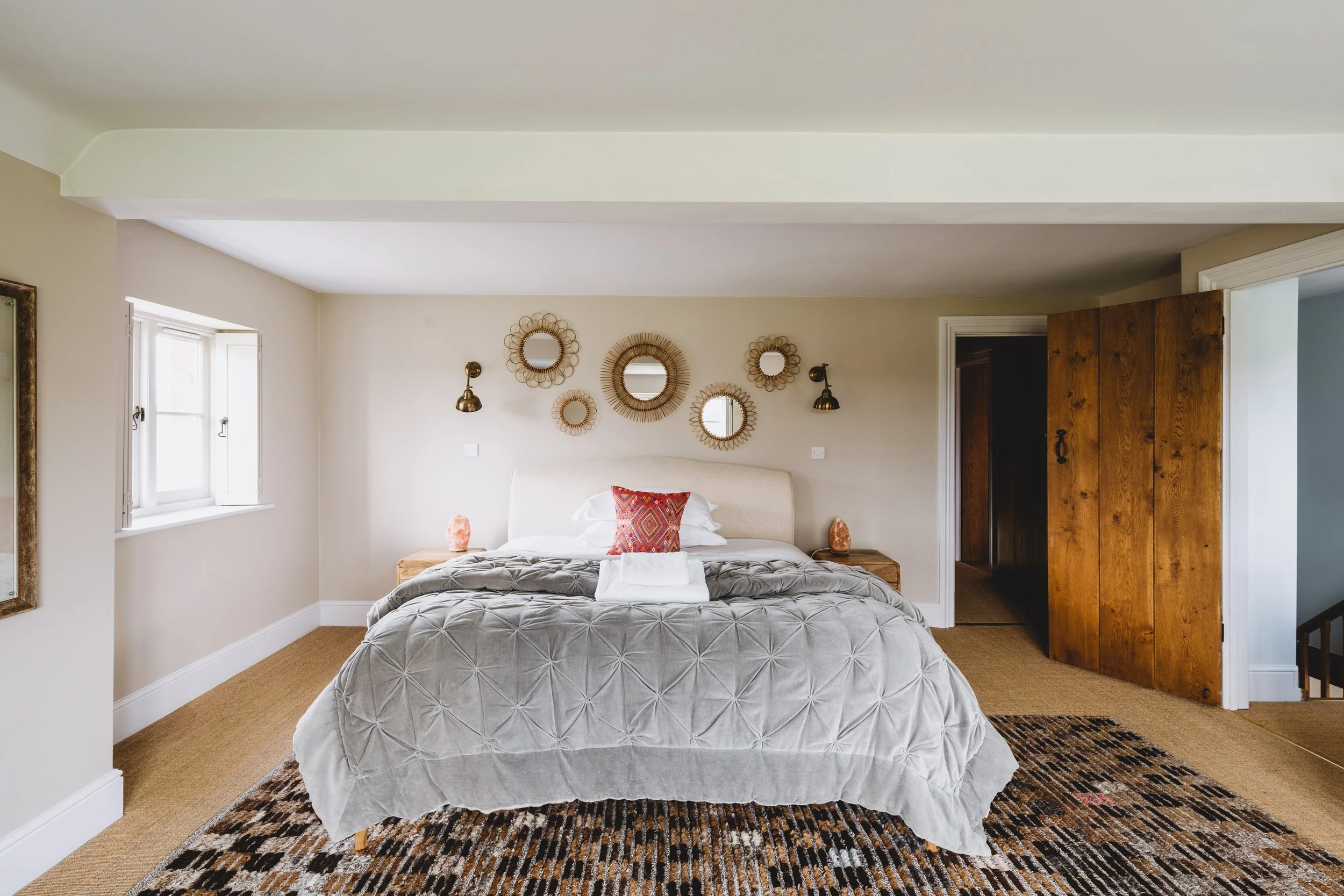 A bedroom with a bed covered in a gray quilt, a colorful decorative pillow, and two salt lamps on wooden nightstands. The wall above the bed has seven circular mirrors and two wall-mounted lamps. There is a window on the left, a wooden door on the right, and a rug on the beige carpeted floor.