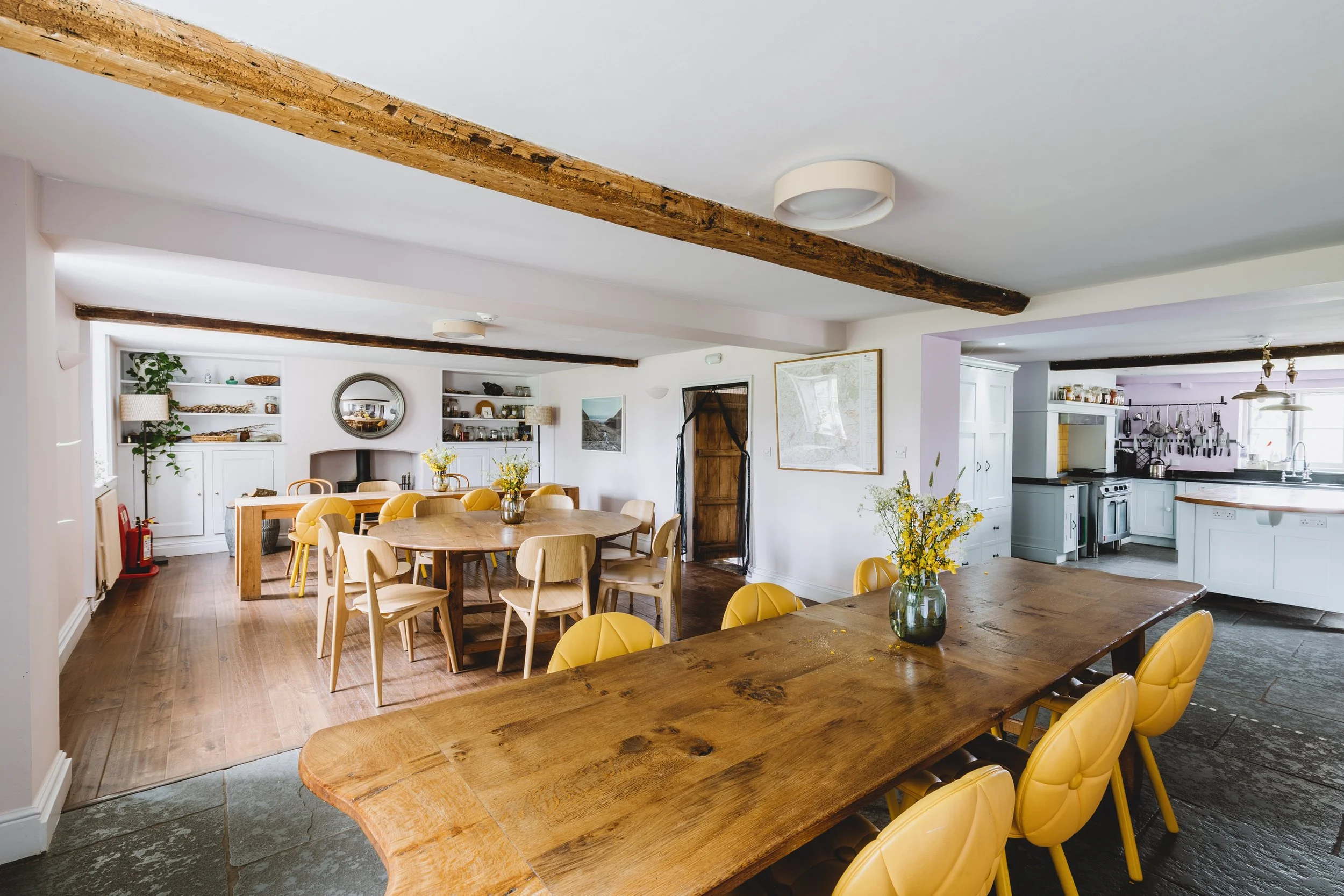 Spacious dining area with wooden tables and yellow chairs, decorated with vases of yellow flowers, adjacent to a kitchen with white cabinets and black countertops.