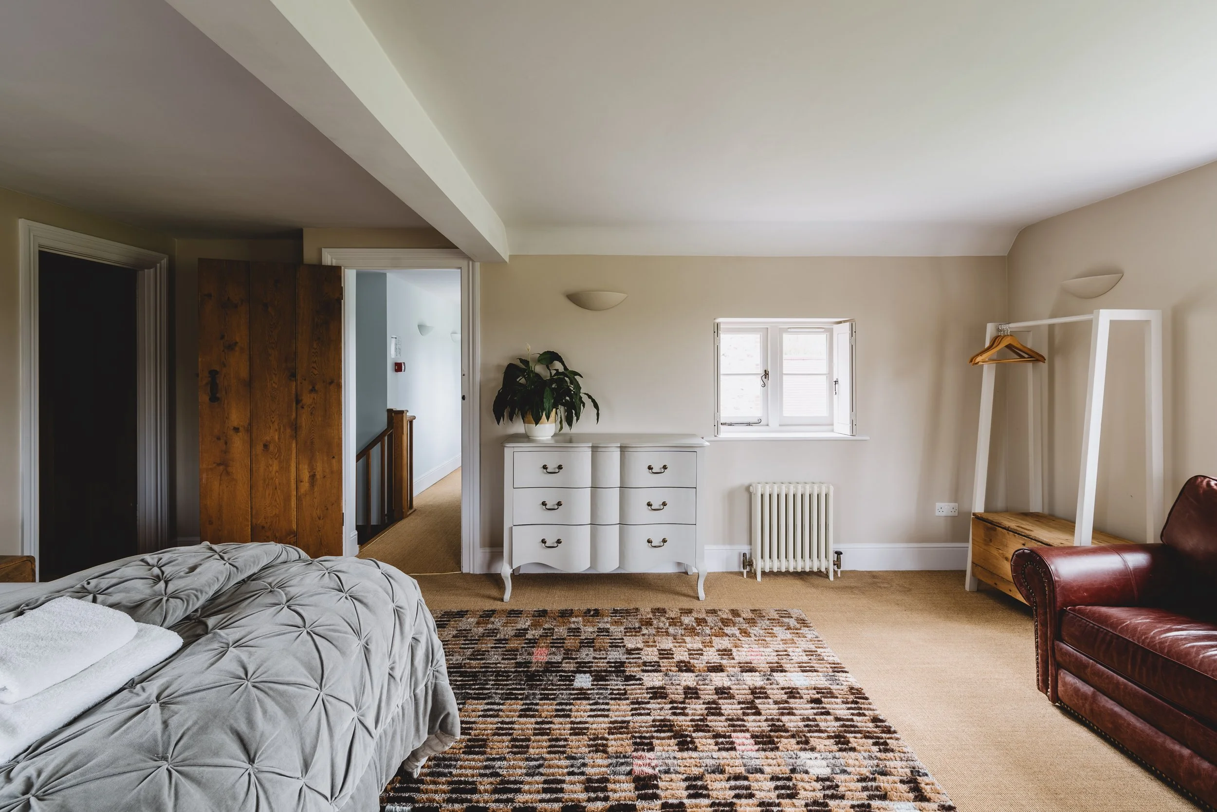 A cozy living room with beige walls and carpet, featuring a bed with a tufted gray blanket, a brown leather sofa, a white dresser, a small window, a decorative woven rug, and a white clothing rack.