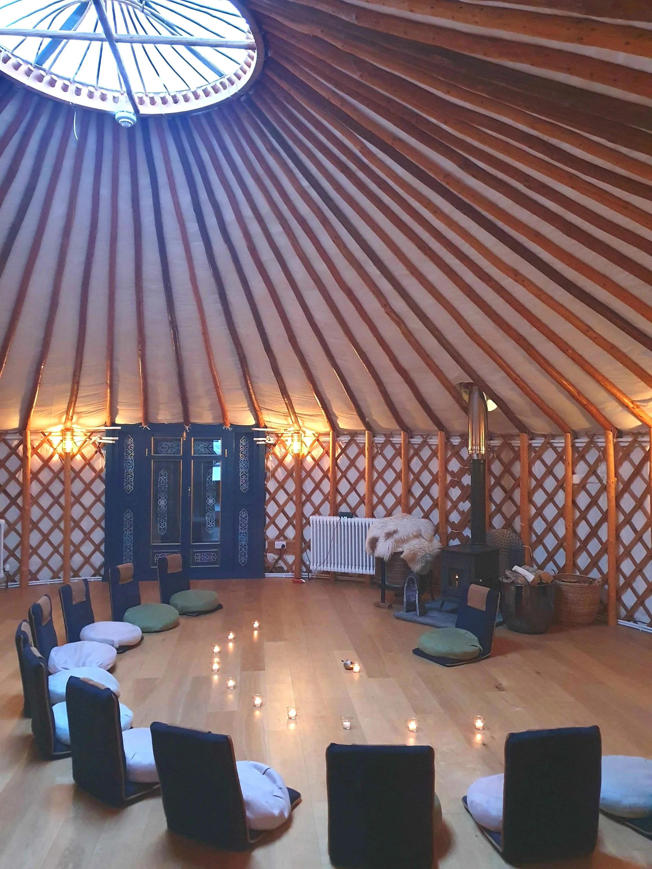 Interior of a yurt with wooden lattice walls and a high, conical ceiling. Chairs with cushions are arranged in a circle on the wooden floor, with small candles placed along the circle. A wood stove, radiator, and decorative door are visible in the background.