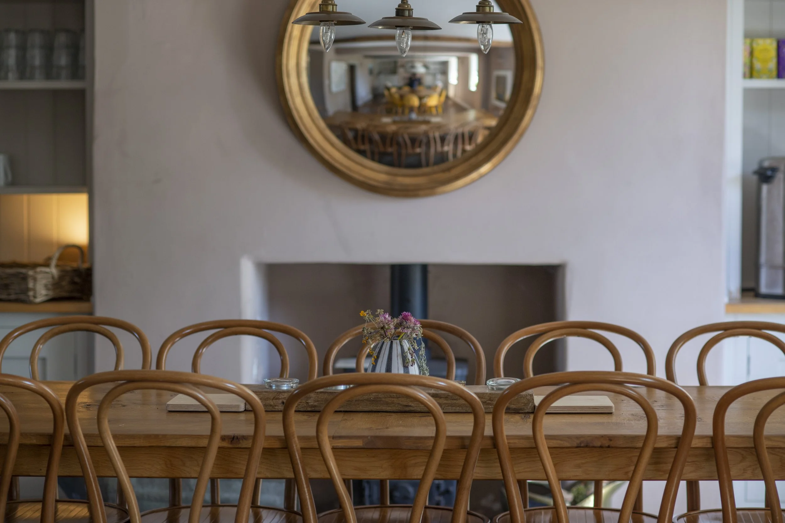 A dining room with a wooden table and matching chairs, a small vase with flowers, a large round mirror on the wall reflecting another room, and a pendant light fixture hanging above the table.