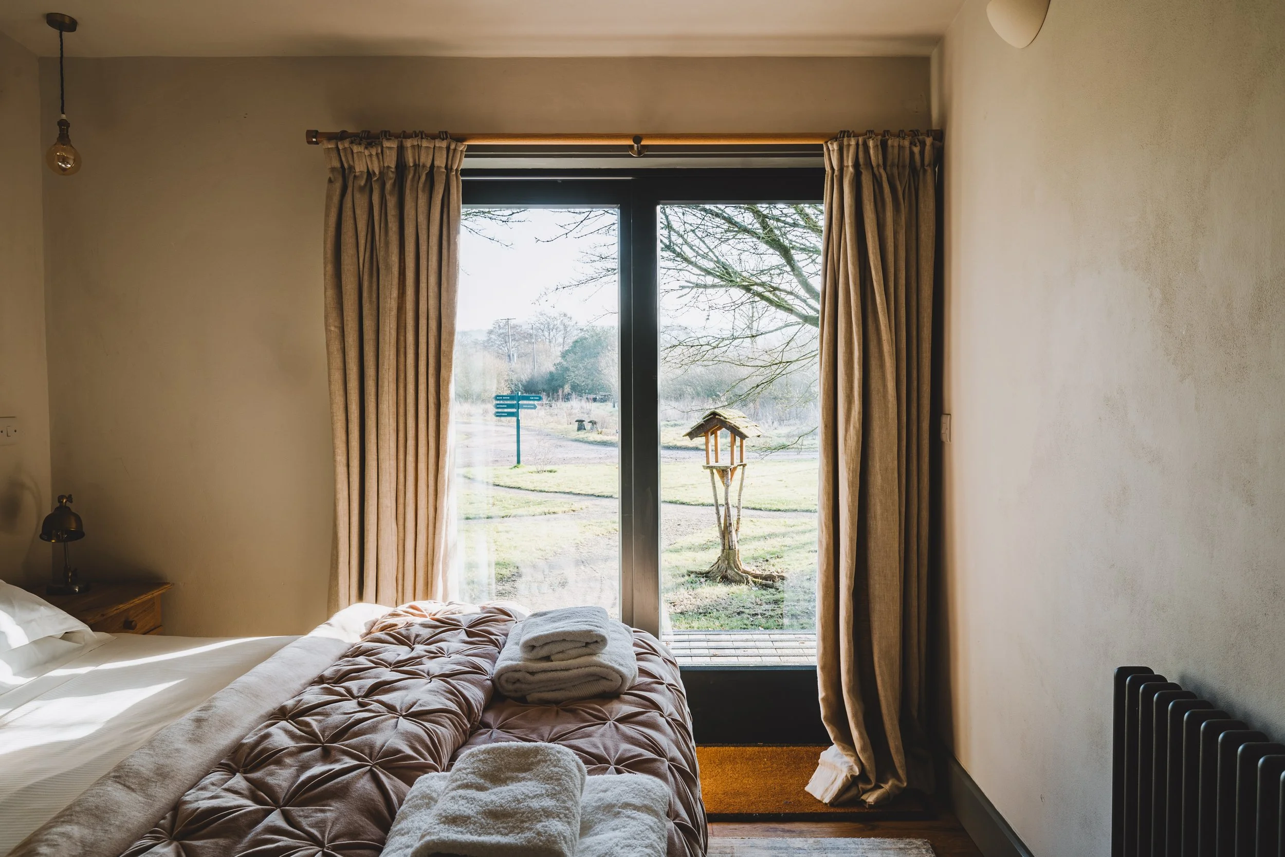 A cozy bedroom with a large window and beige curtains, a bed with rolled towels on top, and a small bedside lamp, overlooking a backyard with a birdhouse and trees.