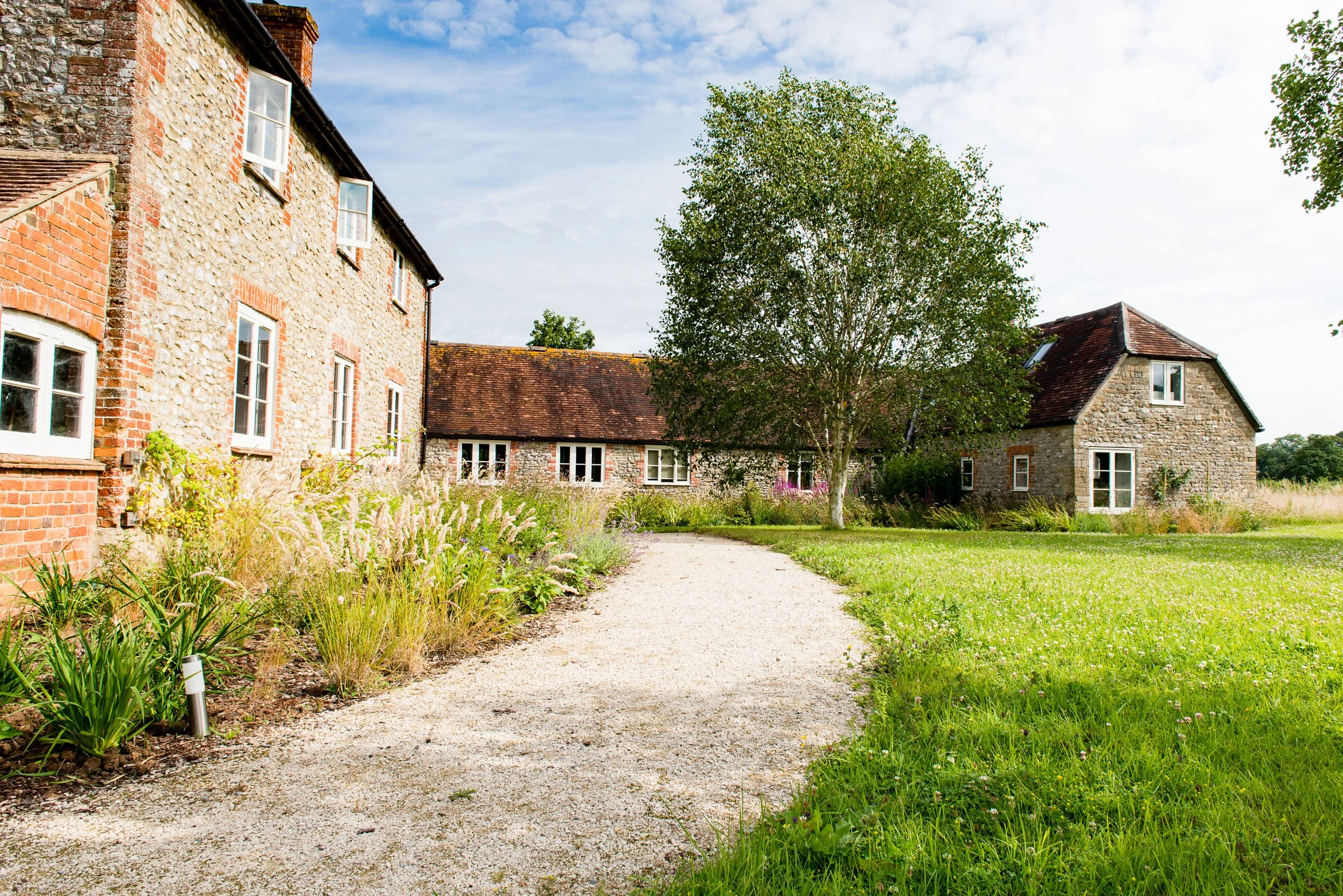 A gravel pathway leads through a lush green garden with grass and flowering plants along the side of a stone and brick building. A large tree with green leaves is in the center, with additional stone buildings with red shingle roofs in the background under a partly cloudy sky.