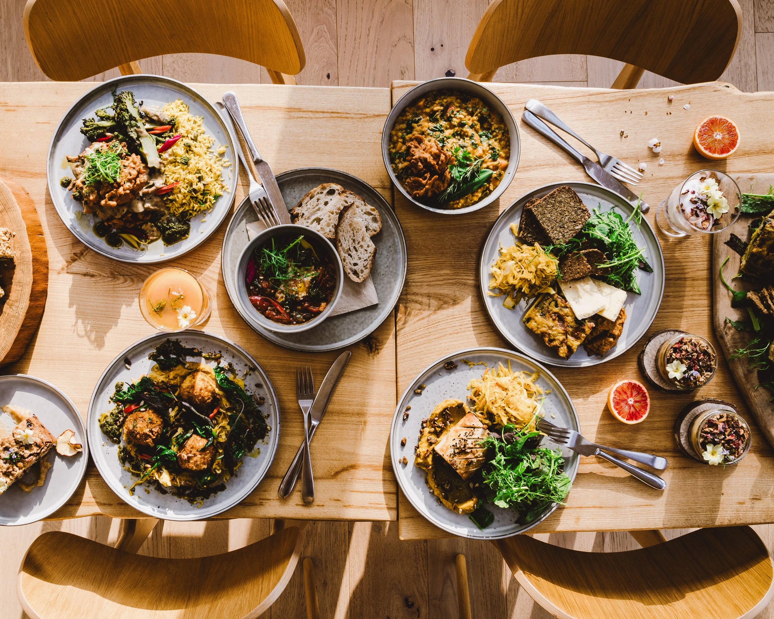 A top-down view of a wooden dining table set with various plates of colorful, diverse dishes, cutlery, bread, and drinks in a cozy, well-lit setting.