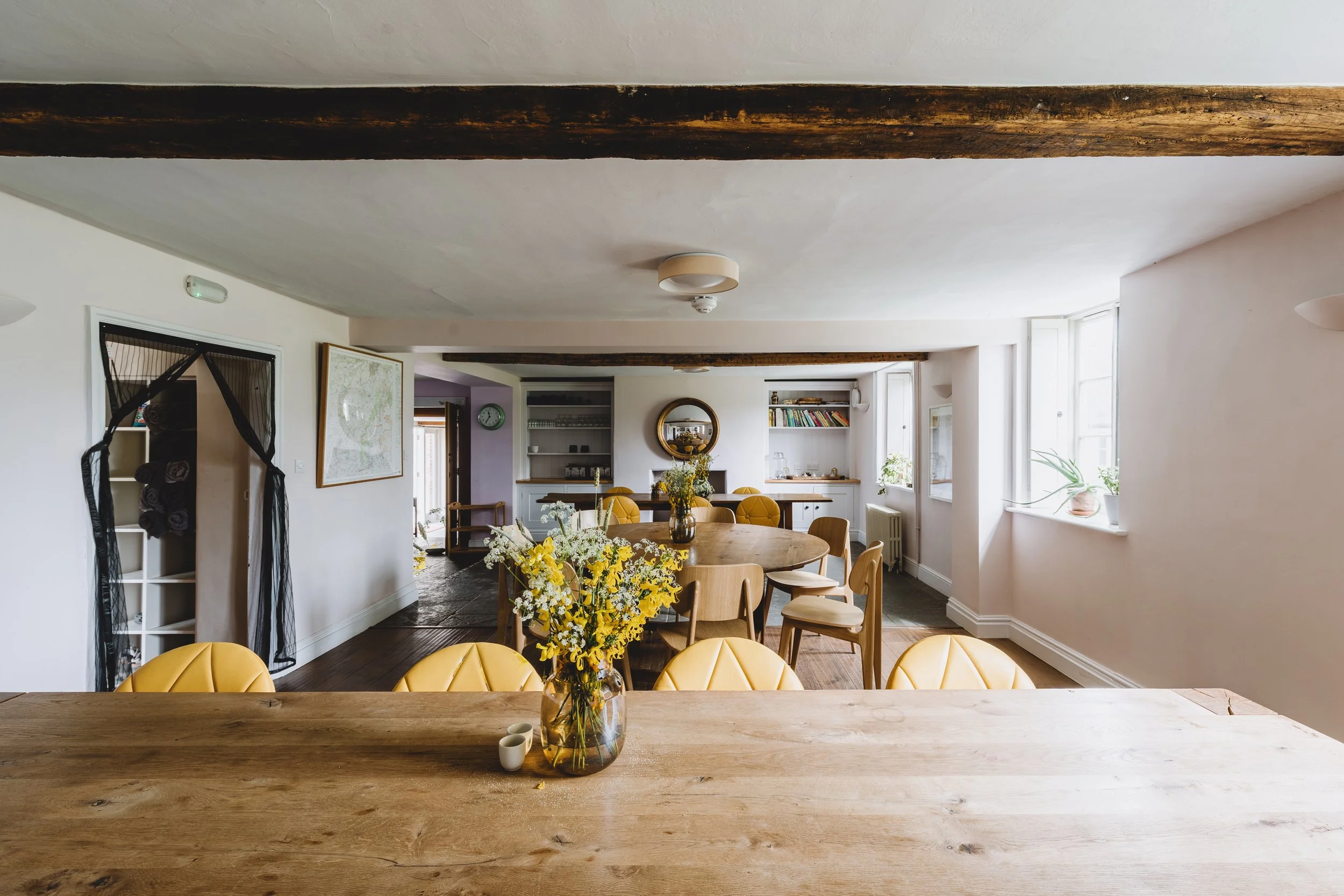 Bright dining area with a wooden table and yellow chairs, a vase with yellow and white flowers, and a round mirror on the wall. Sunlight comes through large windows on the right.