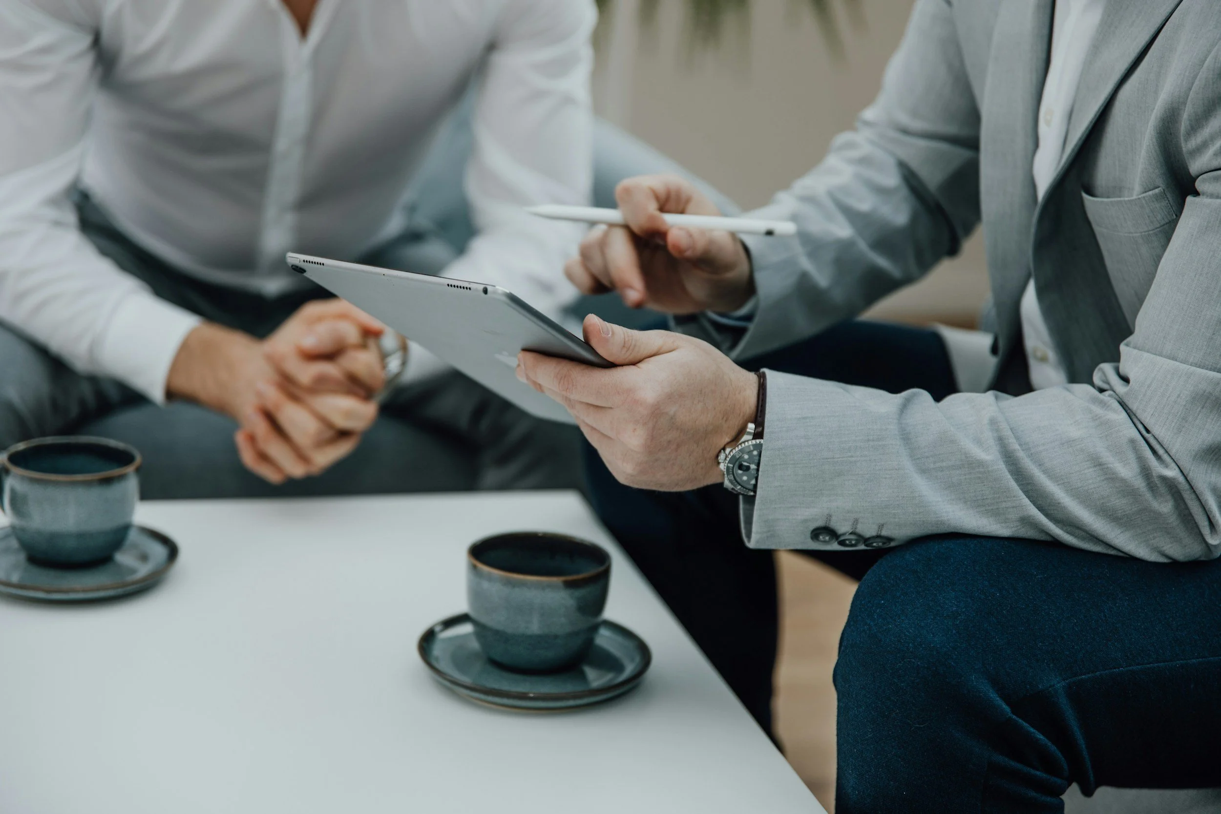 Two men in business attire having a discussion, one is holding a digital tablet and a stylus, the other has clasped hands. There are three cups of coffee on the table.