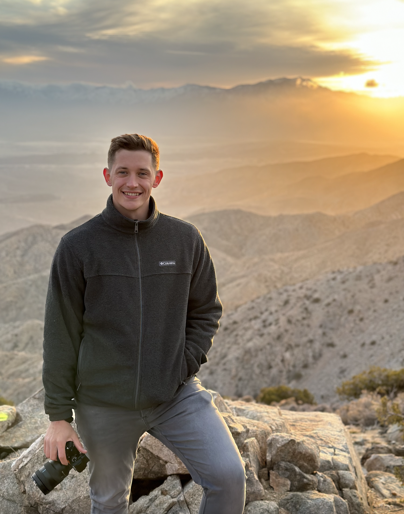 A young man standing on rocky mountainous terrain during sunset, holding a camera and smiling.