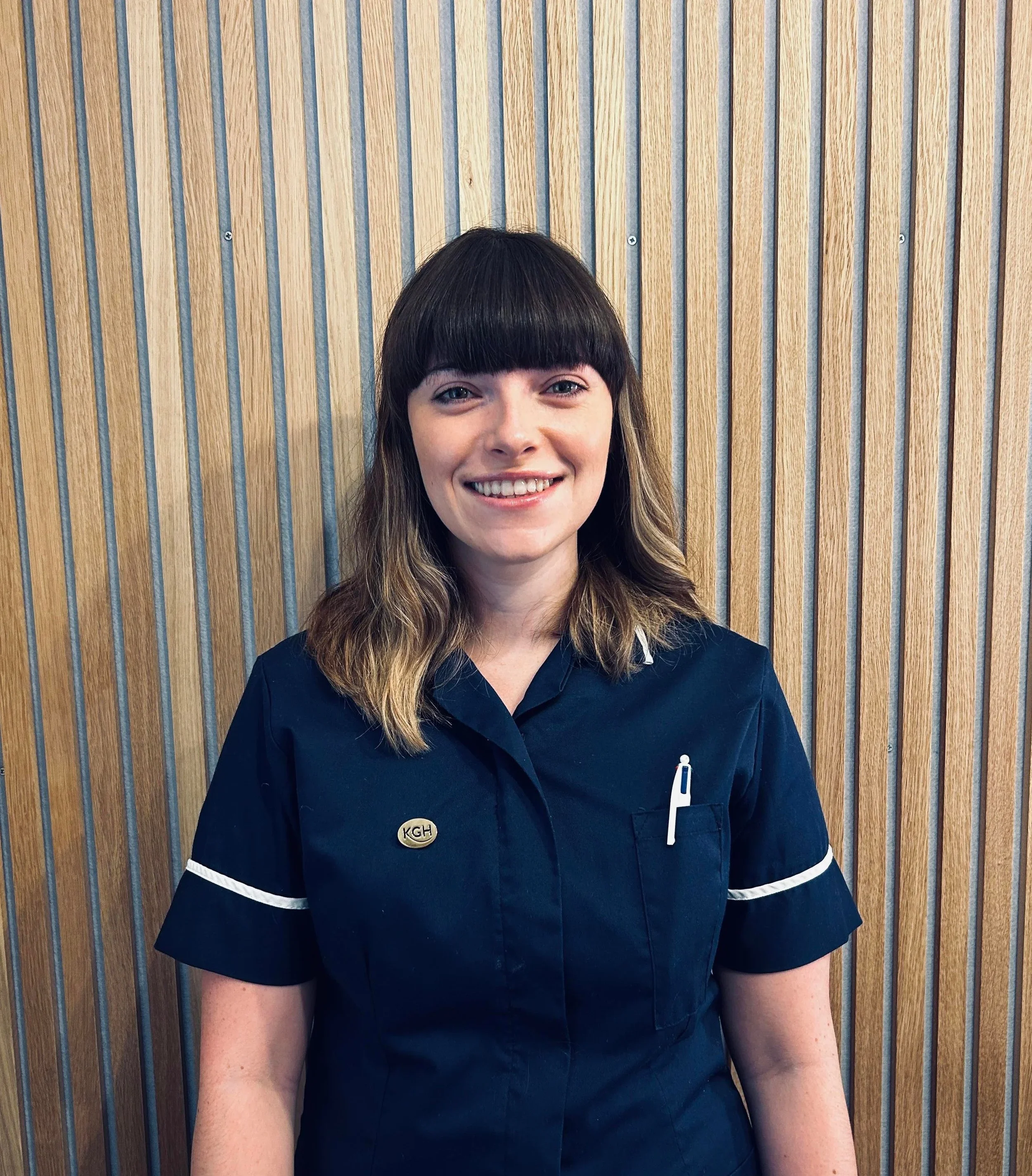 A midwife with shoulder-length brown hair and bangs, smiling, standing in front of a wooden wall, wearing a navy blue outfit with a name badge that reads 'KGH' hypnobirthing and a white pen in her chest pocket.
