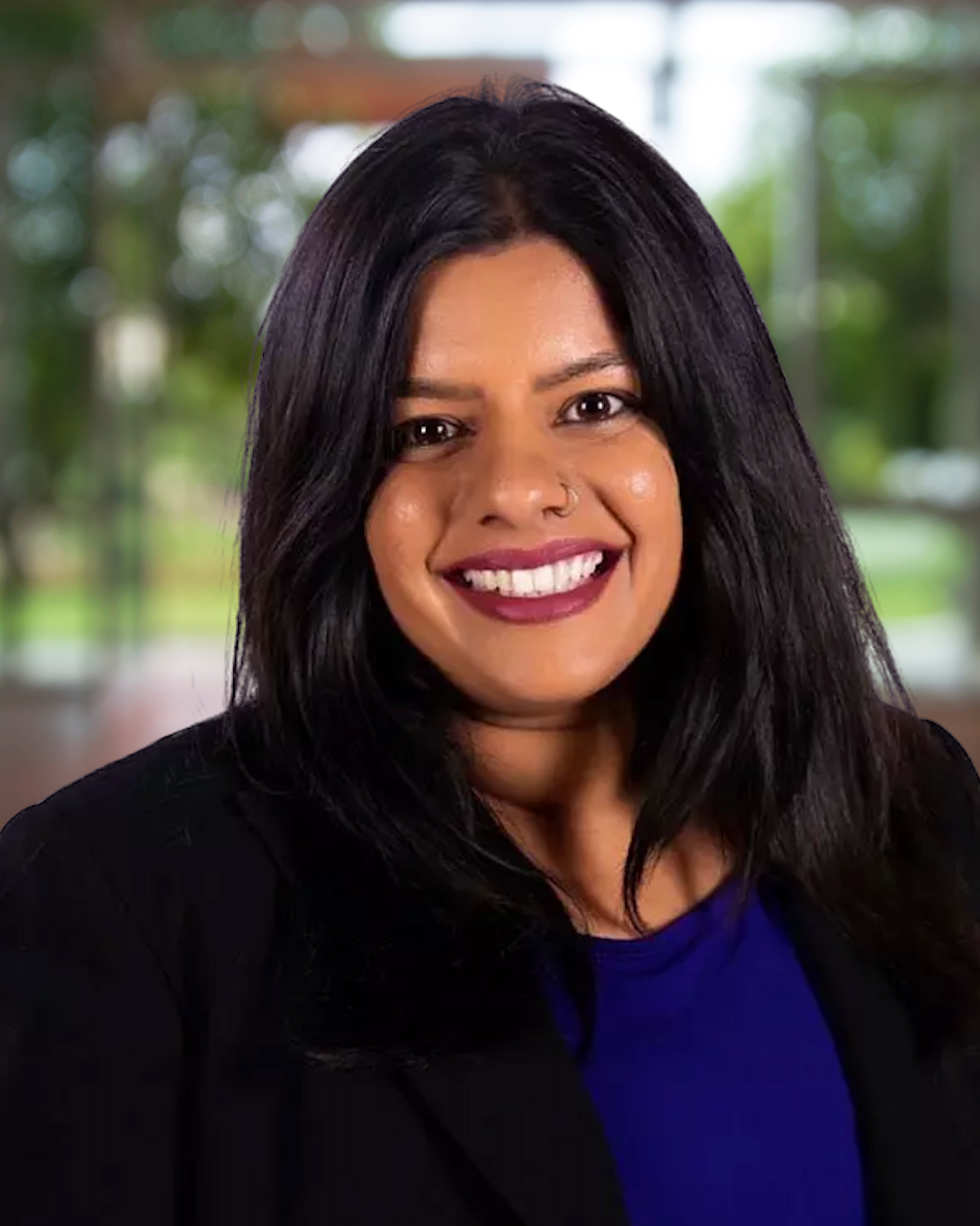 A young woman with long black hair, brown skin, and a nose ring smiling in a professional setting with a blurred background of trees and windows.