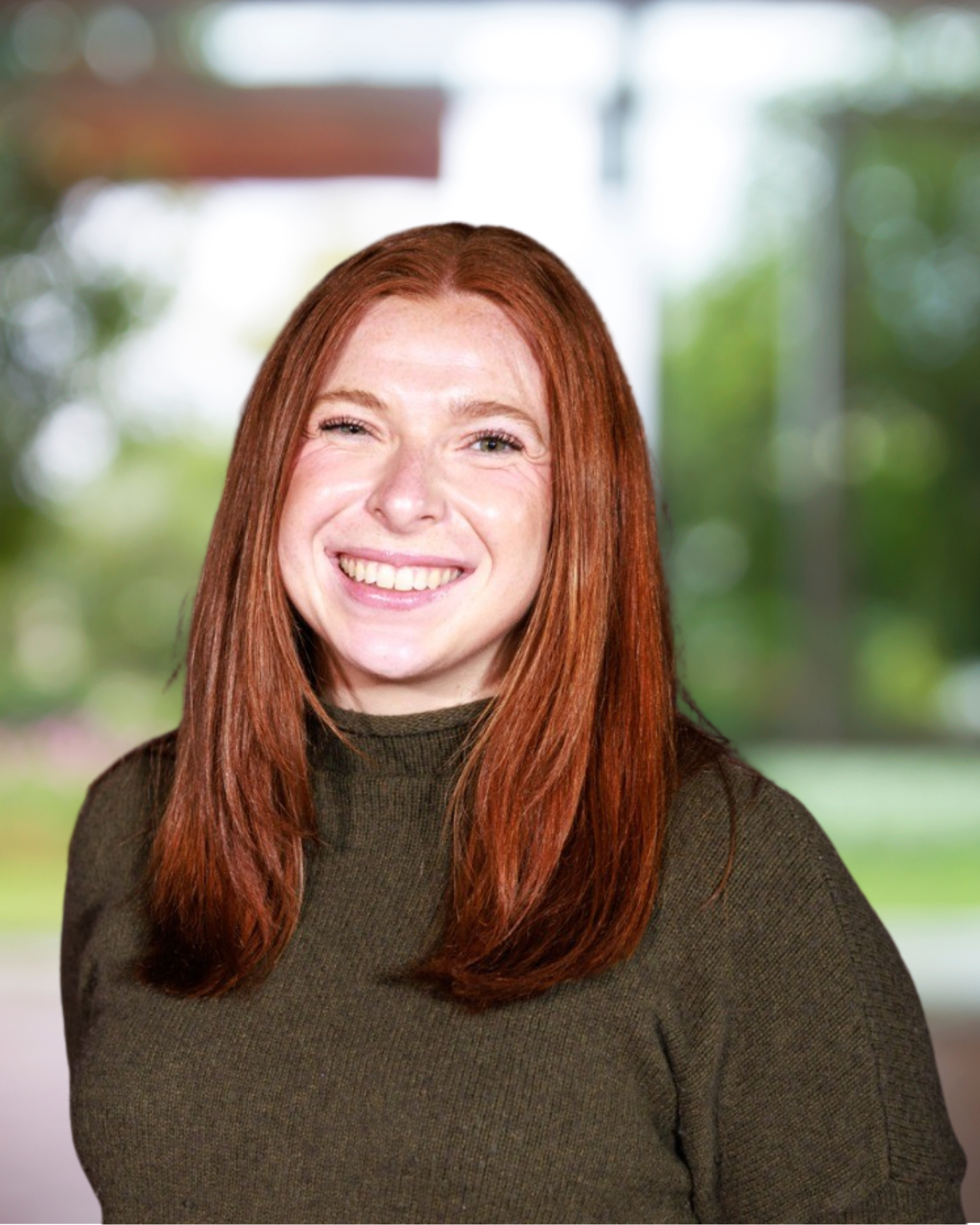 Young woman with red hair smiling indoors with a blurred greenery background.
