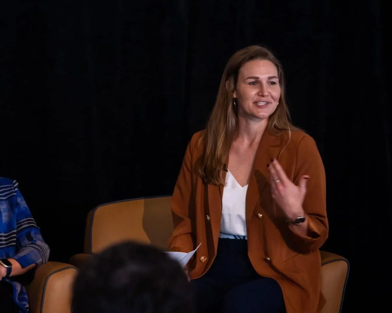 A woman with long brown hair wearing a brown blazer, white blouse, and dark pants, sitting on a beige chair, speaking and gesturing with her hand during a discussion or interview. There are other people partially visible around her.