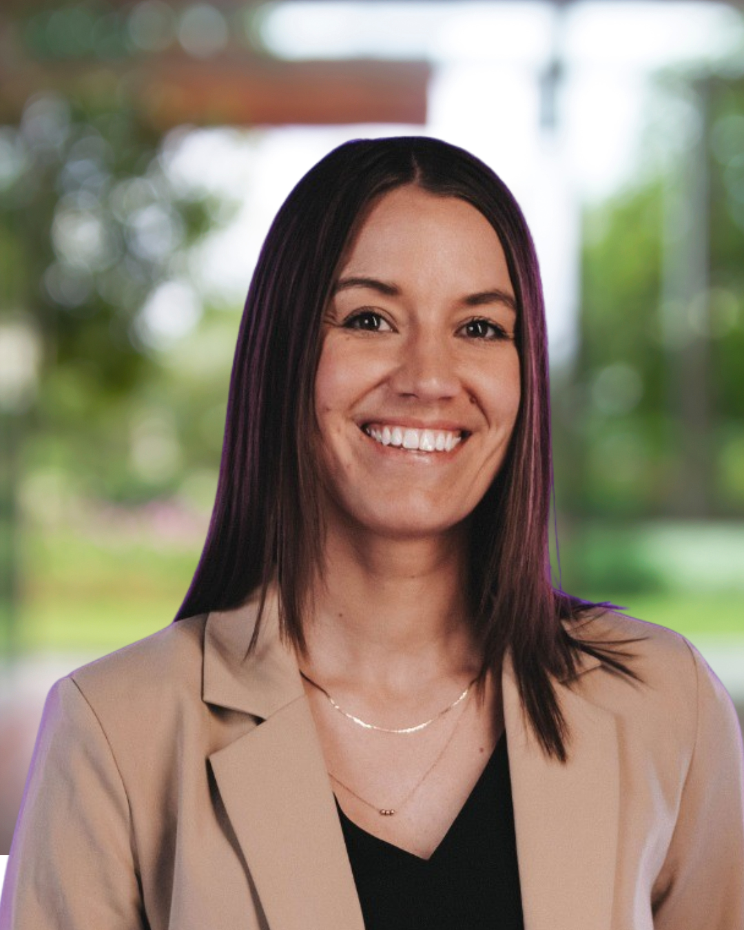 A woman with straight dark brown hair and a big smile, wearing a beige blazer and black top, standing outdoors with greenery in the background.
