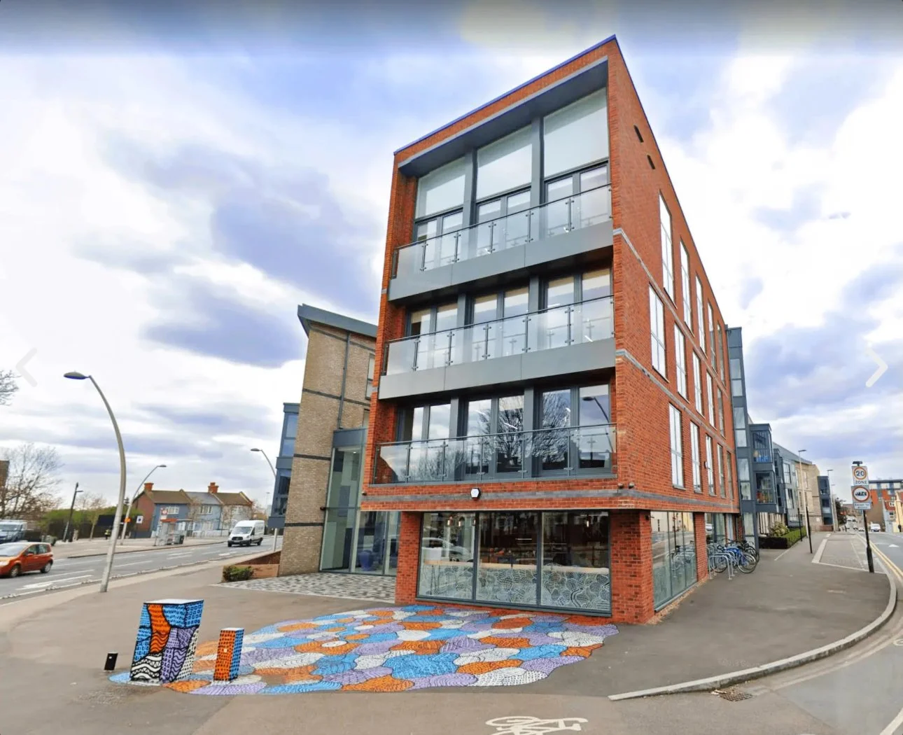 Modern four-story building with brick and glass exterior, balconies on each floor, bicycle parking, colorful mosaic sidewalk in front, and a street with cars and speed limit signs.