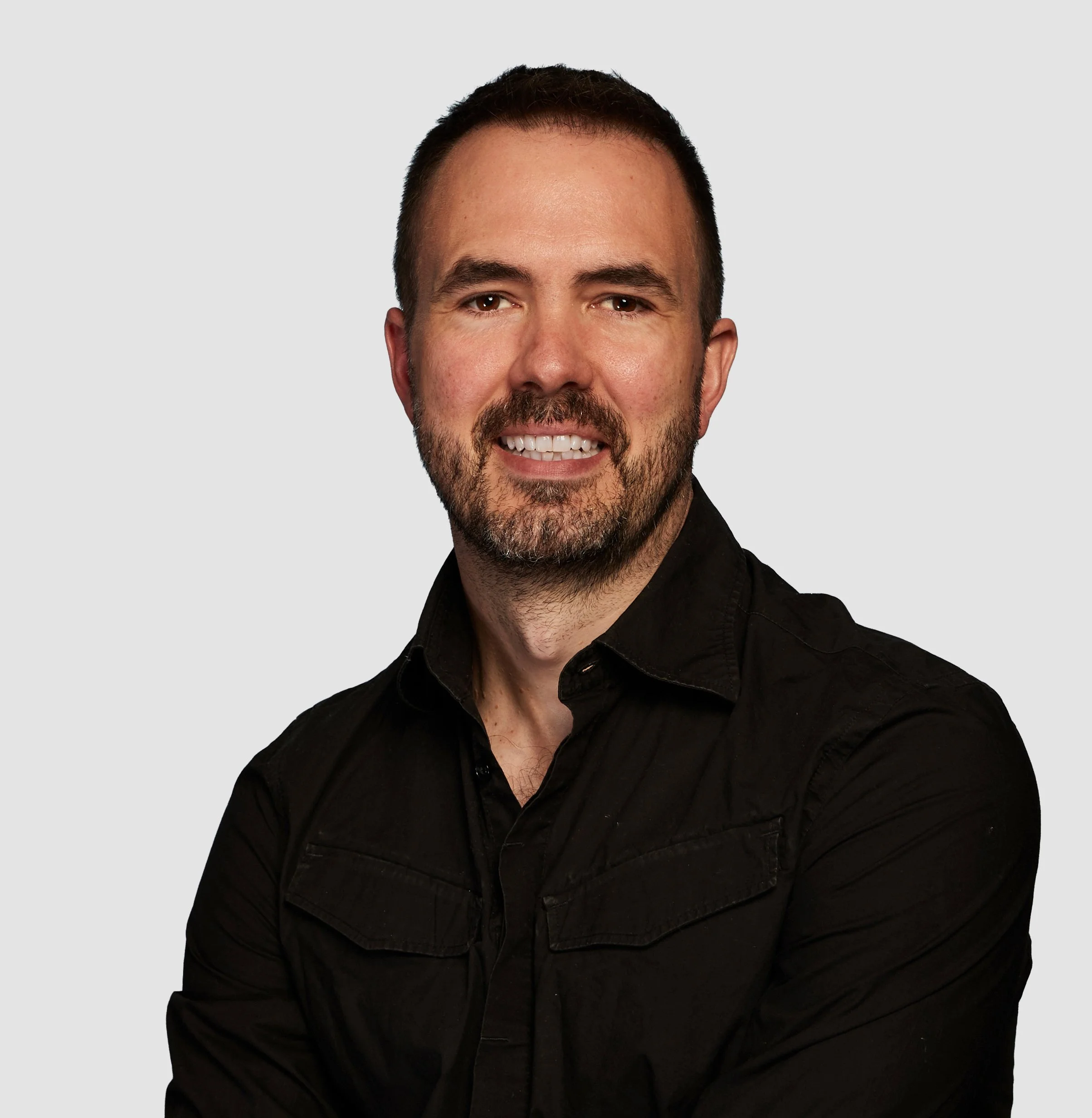 A man with short dark hair, a beard, and a friendly smile wearing a black shirt, against a plain light gray background.