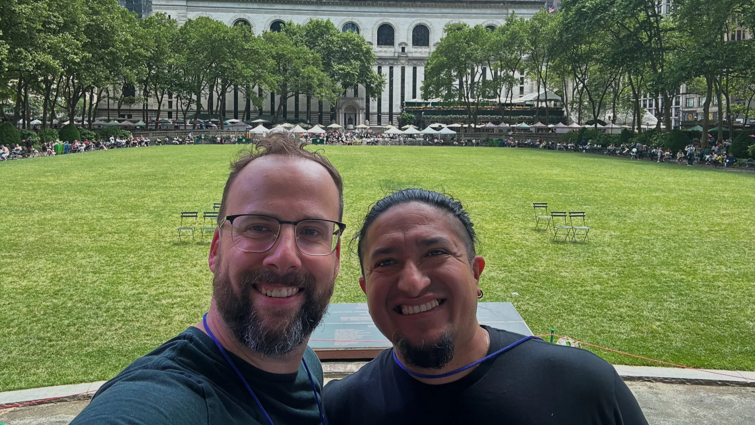 Two men taking a selfie in front of a green park with chairs, trees, and a large historical building in the background.