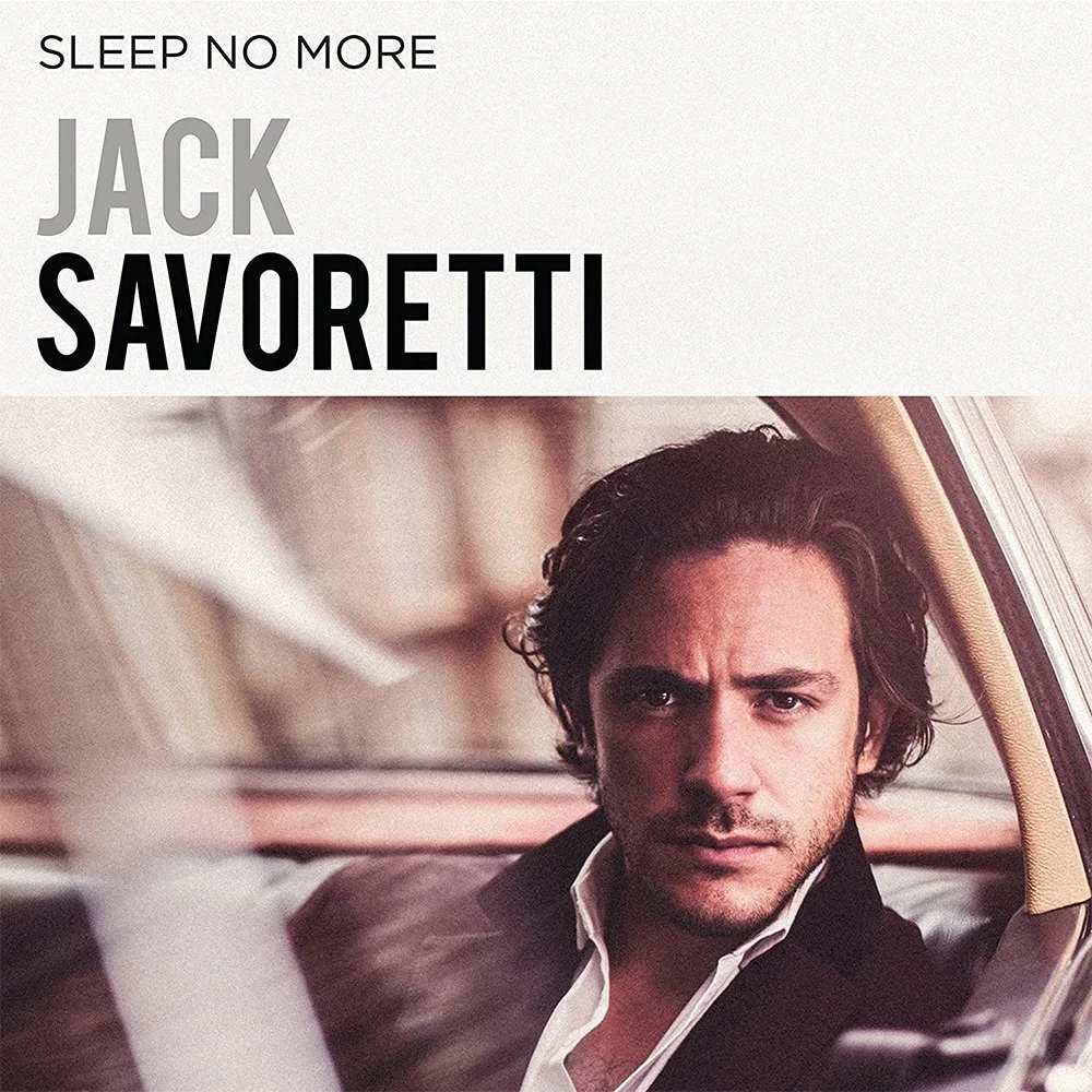 Album cover for Jack Savoretti's "Sleep No More" featuring a close-up of a man with dark, wavy hair, dressed in a black jacket and white shirt, sitting in a car with serious expression.