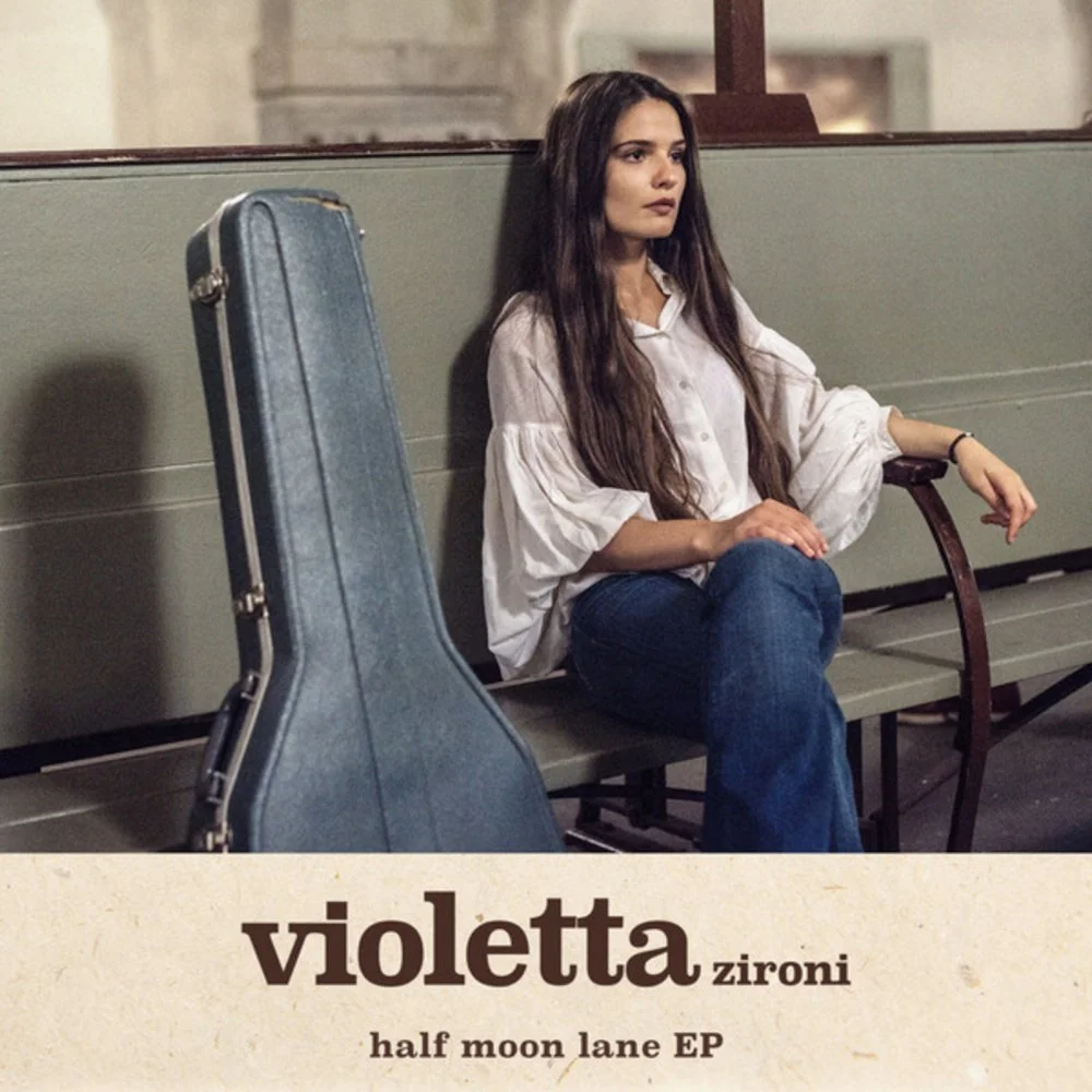 A young woman with long dark hair sitting alone on a bench in a waiting area, with a guitar case leaning against the bench next to her. She is wearing a white blouse and jeans, looking contemplative.