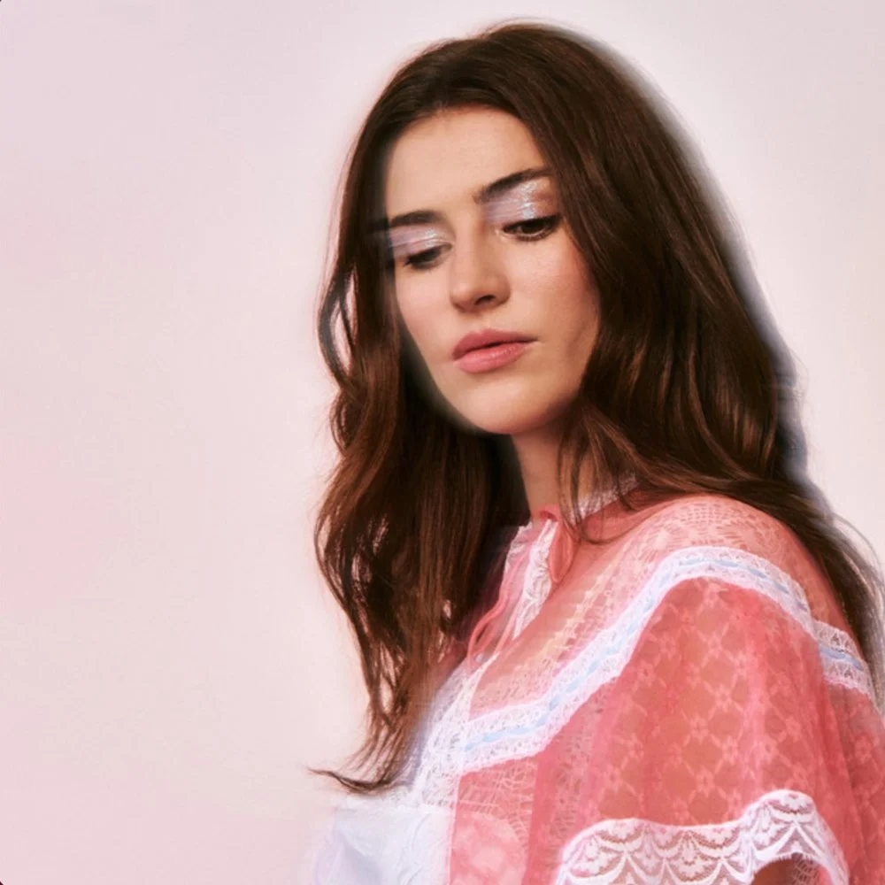 A woman with long brown hair and light makeup, wearing a pink and white lace dress, looking down with a soft expression.