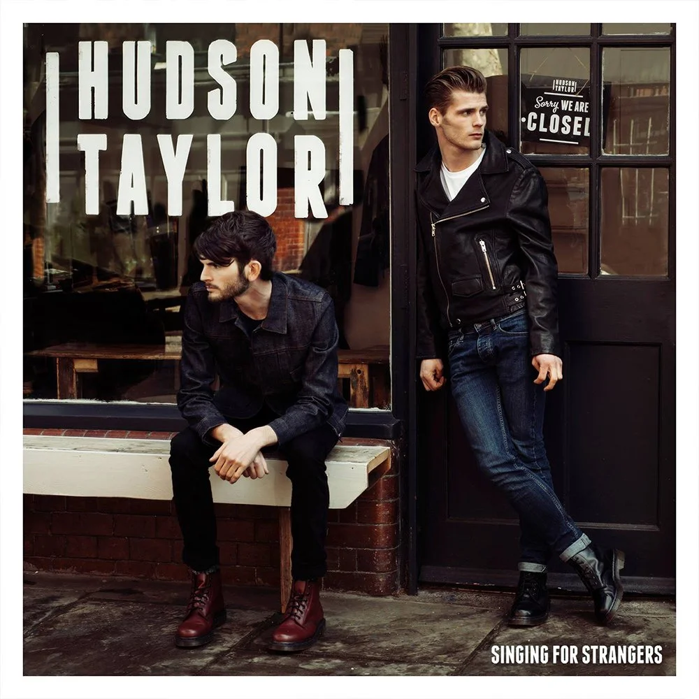 Two young men in dark clothing outside a store with a sign that reads "Hudson, Taylor" and a window with a sign that says "Sorry we are closed". One is sitting on a bench, and the other is standing near the door, looking to the side.