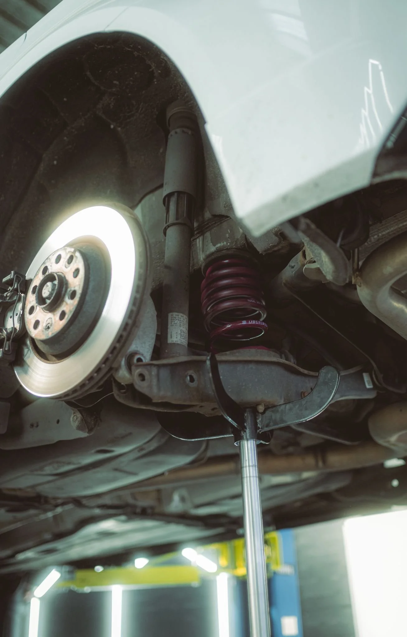 Underneath view of a car suspension system with a coil spring, shock absorber, brake disc, and parts of the steering mechanism visible.