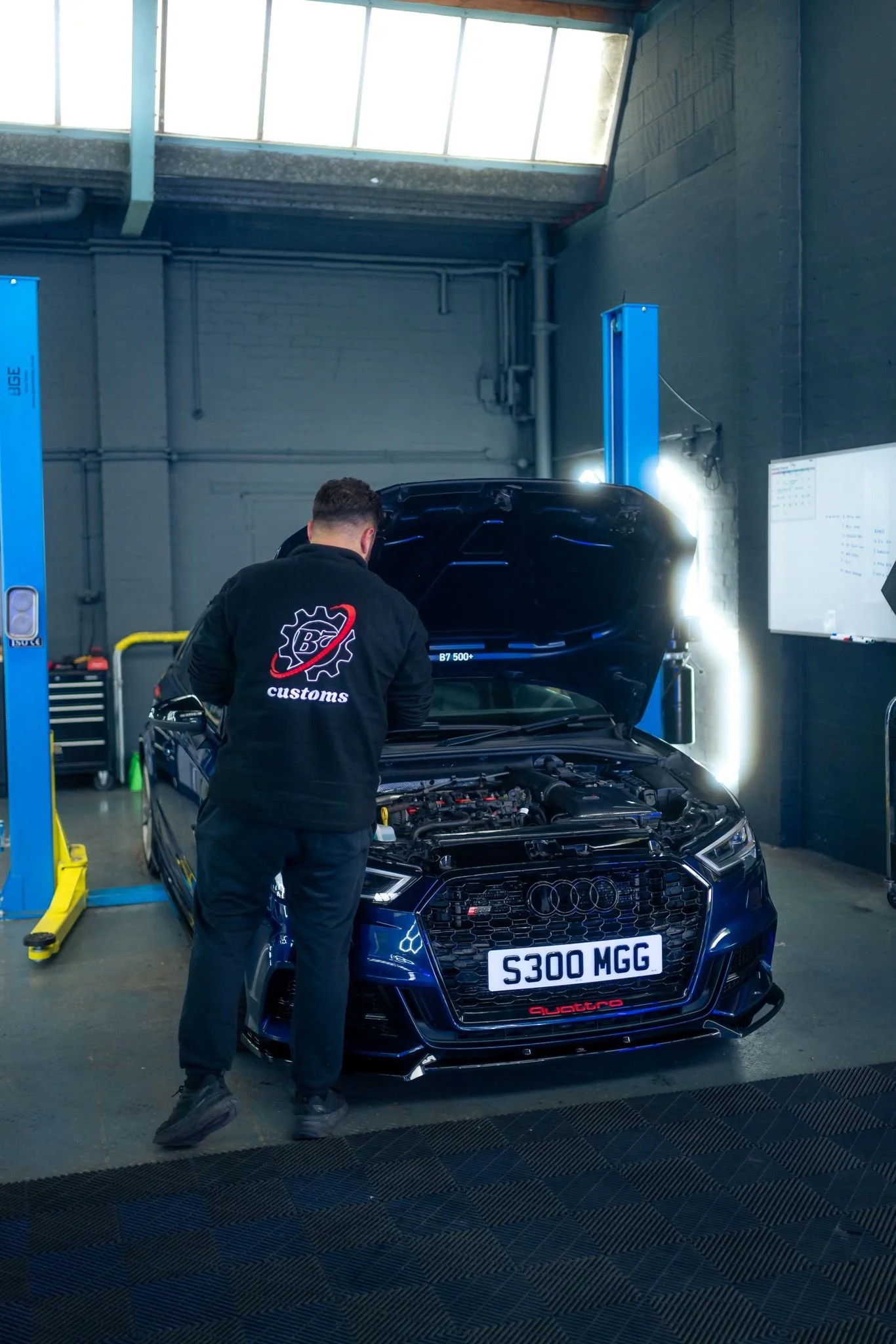A mechanic working on a black Audi car with an open hood inside a garage or workshop, with tools and equipment visible in the background.