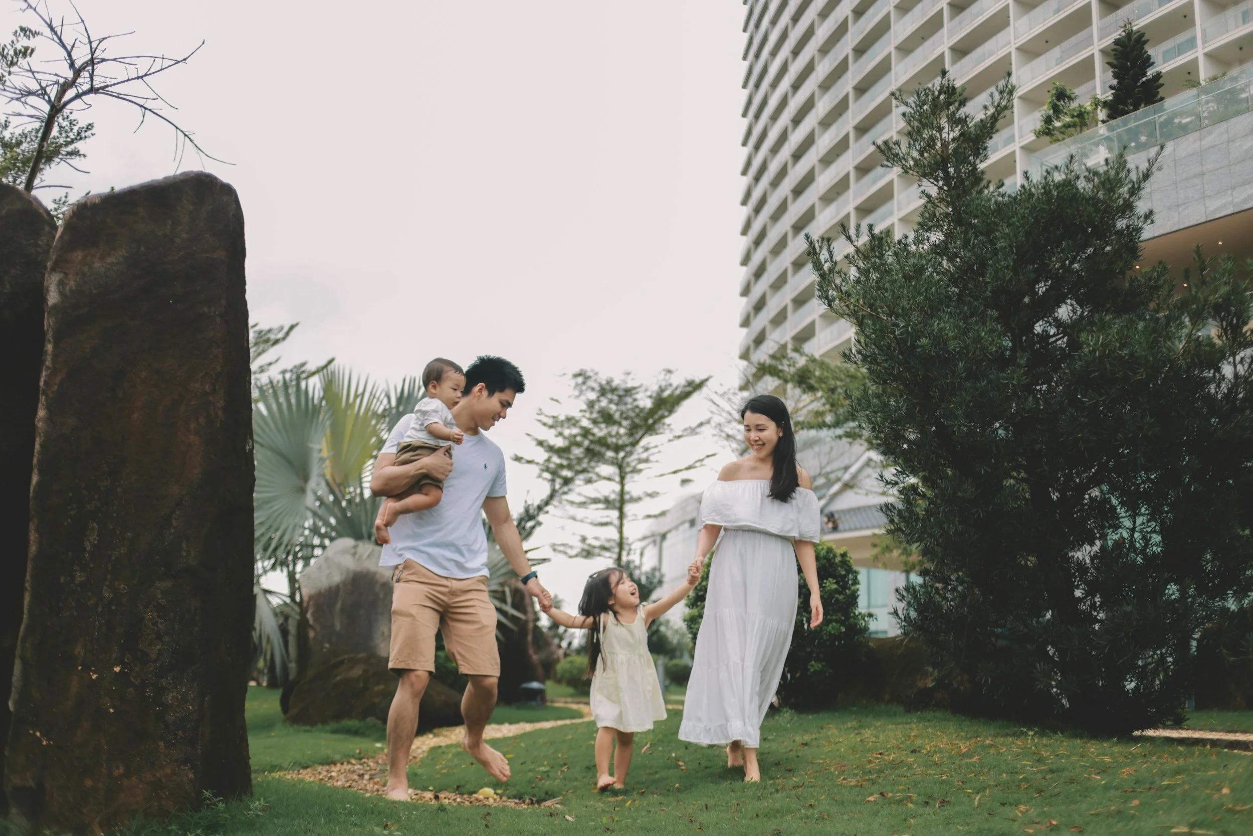 A family of four playing outside on a grassy lawn, holding hands and smiling, with a modern building and trees in the background.