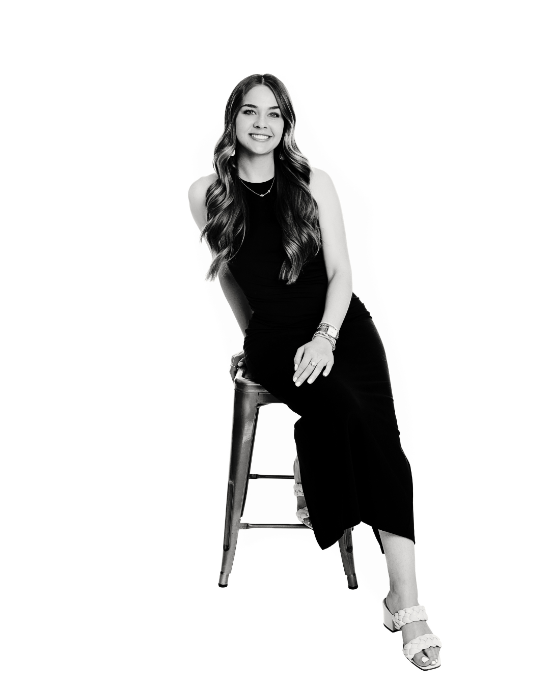 Ireland is sitting on a stool, smiling, with long wavy hair, wearing a sleeveless dress, jewelry, and sandals, against a plain background.