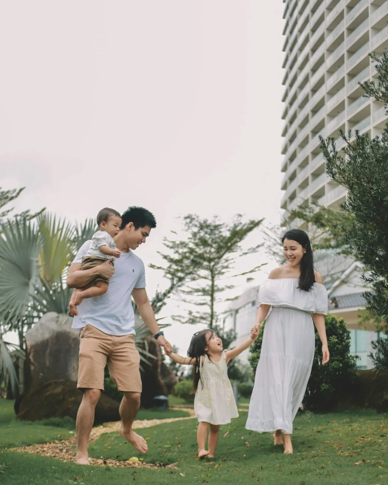 A family of four playing outside on a grassy lawn, holding hands and smiling, with a modern building and trees in the background.