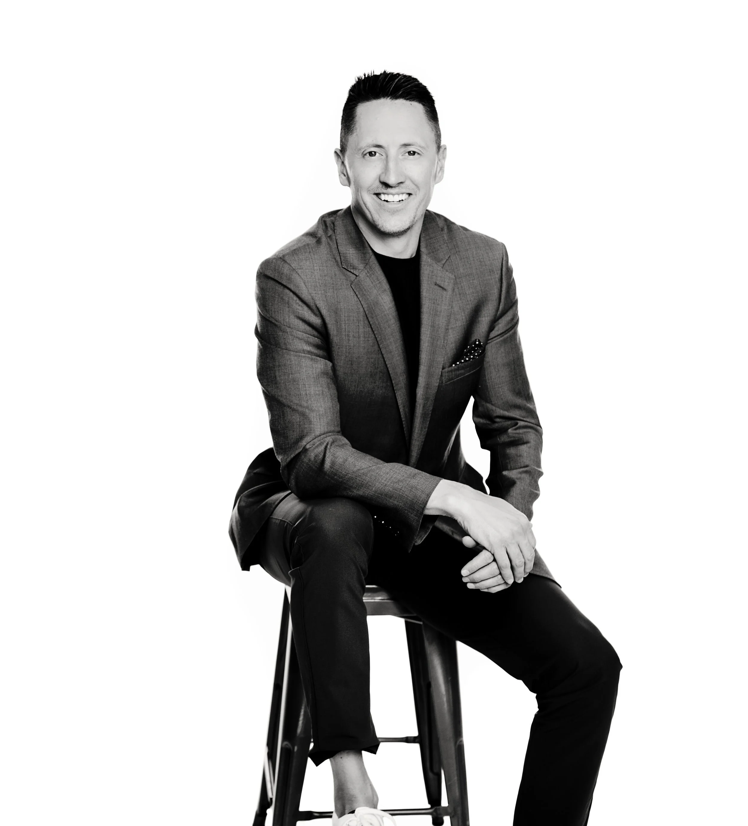 Keiven is dressed in a suit, sitting on a stool against a white background.