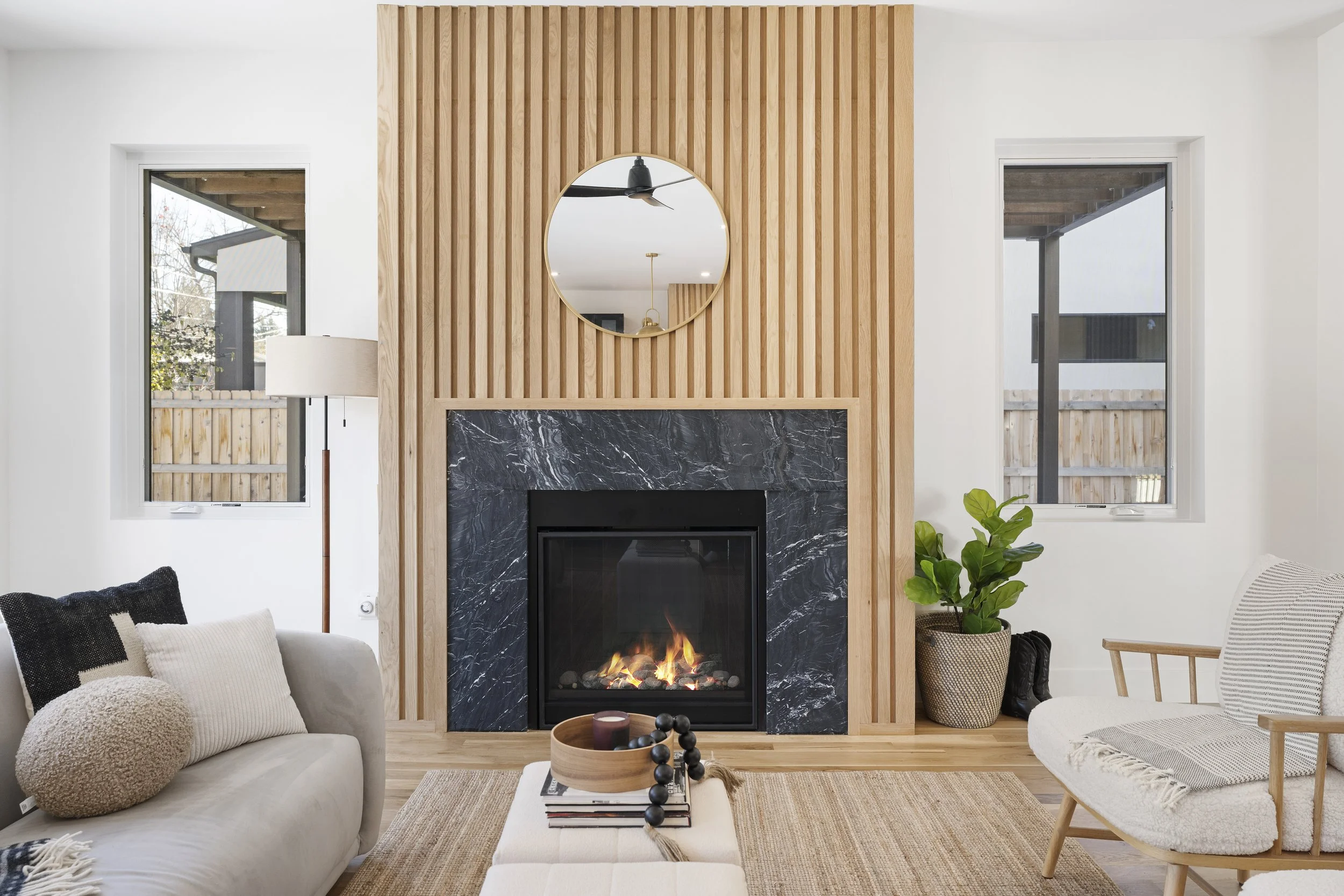 Living room with a modern fireplace with black marble surround, light wood panel wall, round mirror above the fireplace, beige sofa on the left, wooden chair with woven seat on the right, large potted plant, two windows, and a coffee table with decorative items.