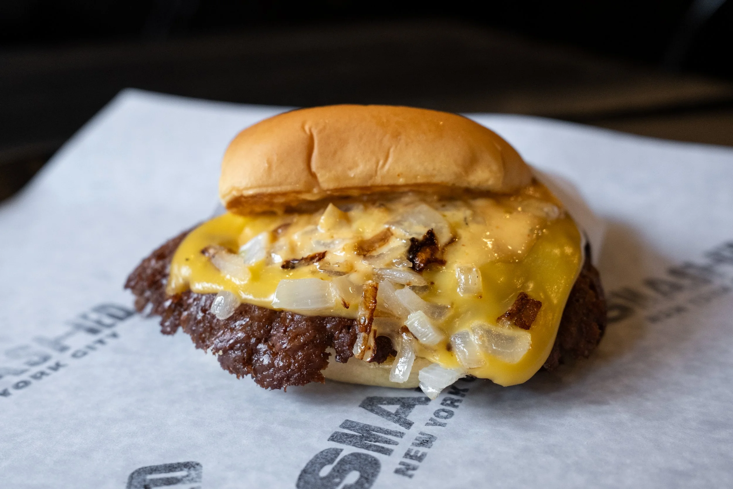 Close-up of a cheeseburger with a beef patty, melted cheese, grilled onions, and a small sandwich bun on top, placed on white paper.