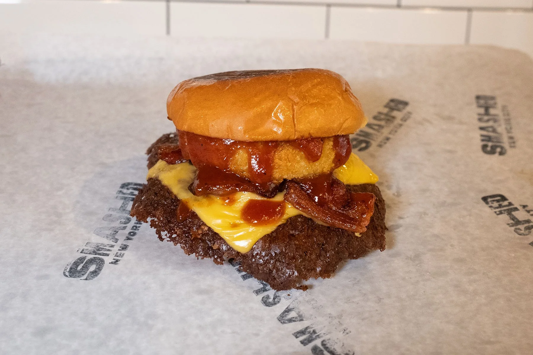 Close-up of a burger with a fried chicken fillet, melted cheese, bacon, barbecue sauce, and a sandwich bun on top, on a piece of branded paper.