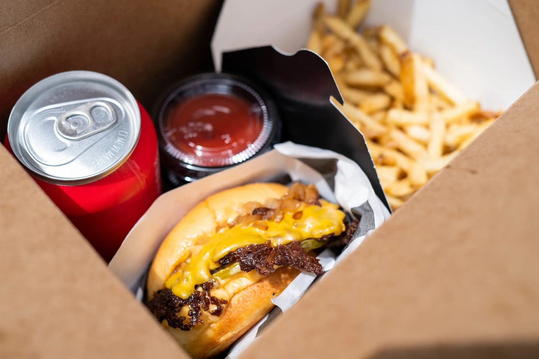 A takeout box containing a cheeseburger with bacon, cheddar cheese, and toppings, French fries, a can of soda, and a small container of ketchup.