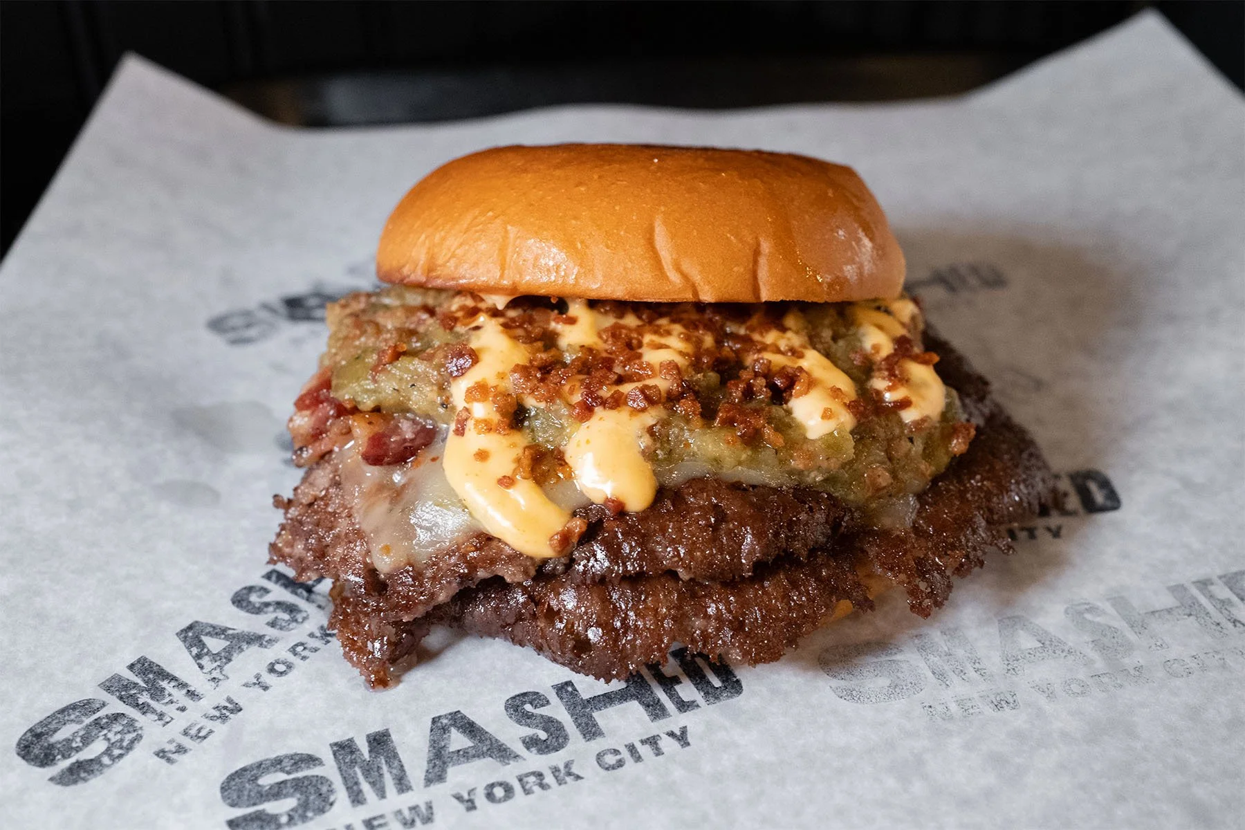 Close-up of a smashed burger with toppings on branded parchment paper, on a dark surface.