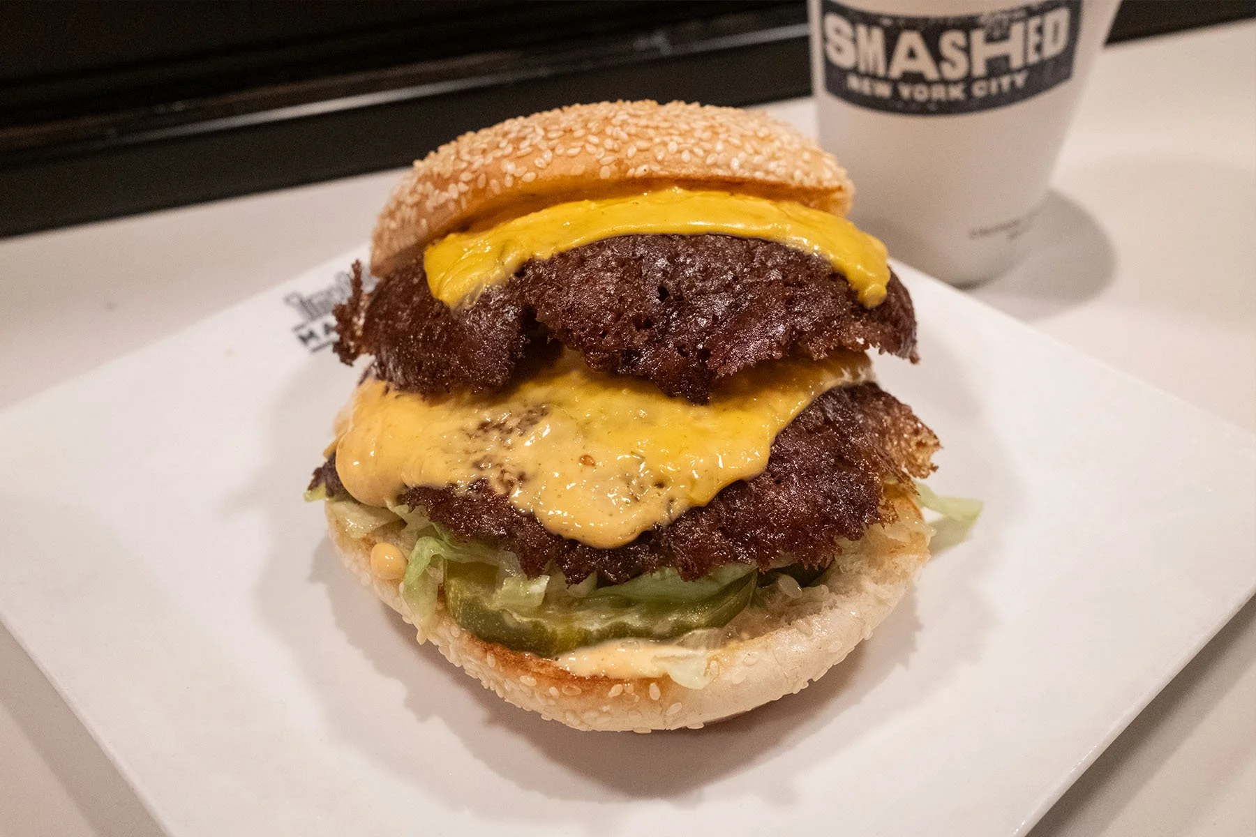 A cheeseburger with two beef patties, melted cheese, pickles, lettuce, and a sesame seed bun, served on a white tray next to a paper cup with the logo 'SMASHED NEW YORK CITY'.